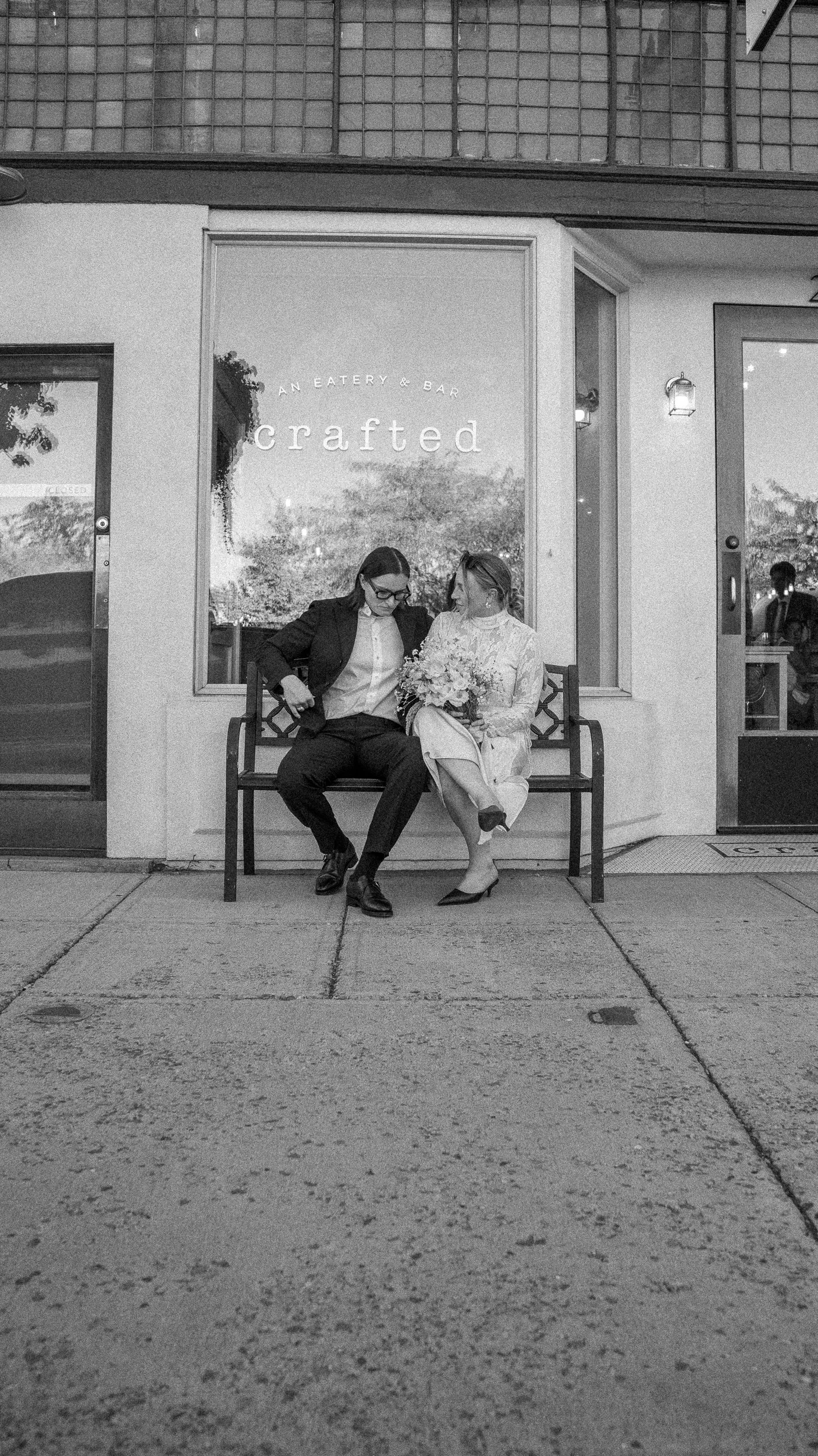 Two women sitting on a bench in front of a restaurant, one holding a bouquet of flowers, the other adjusting her blazer, with the restaurant sign 'crafted' behind them