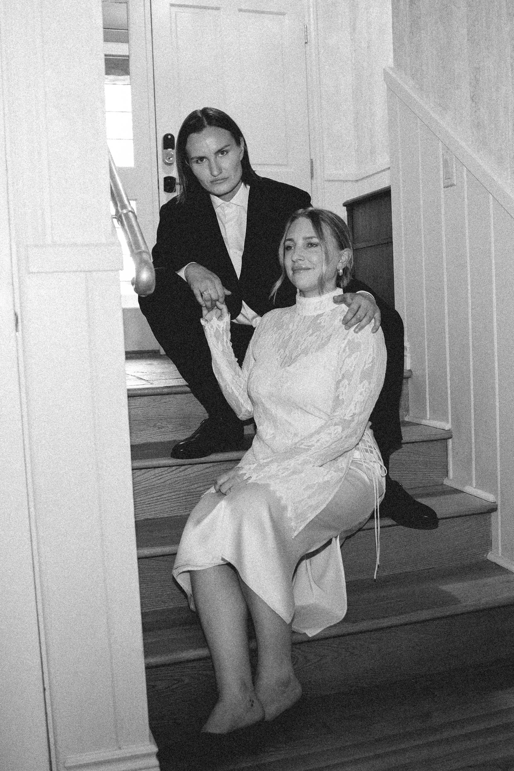 A black-and-white photo of two women on indoor stairs. The woman sitting on the bottom step is dressed in a lace top and satin skirt, with short hair and a pleasant expression. The woman standing on the stairs behind her is dressed in a dark suit, wi