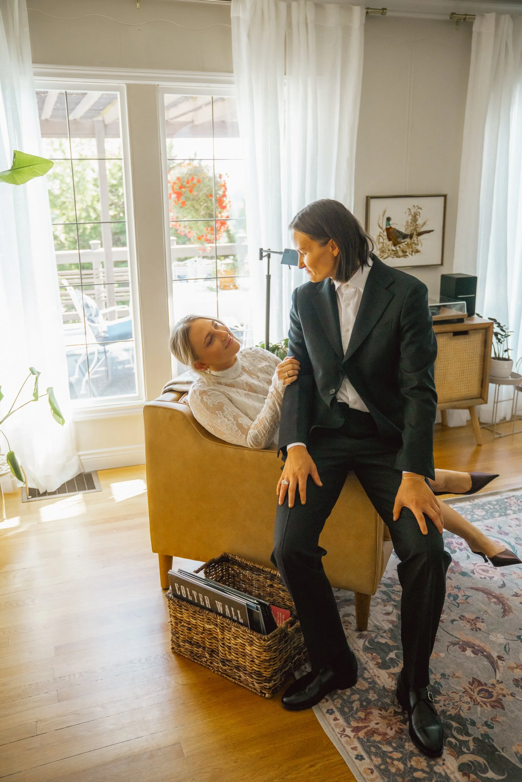 Woman sitting on a sofa, looking up at a woman in a black suit who is standing close to her, with a bright room and large windows in the background.