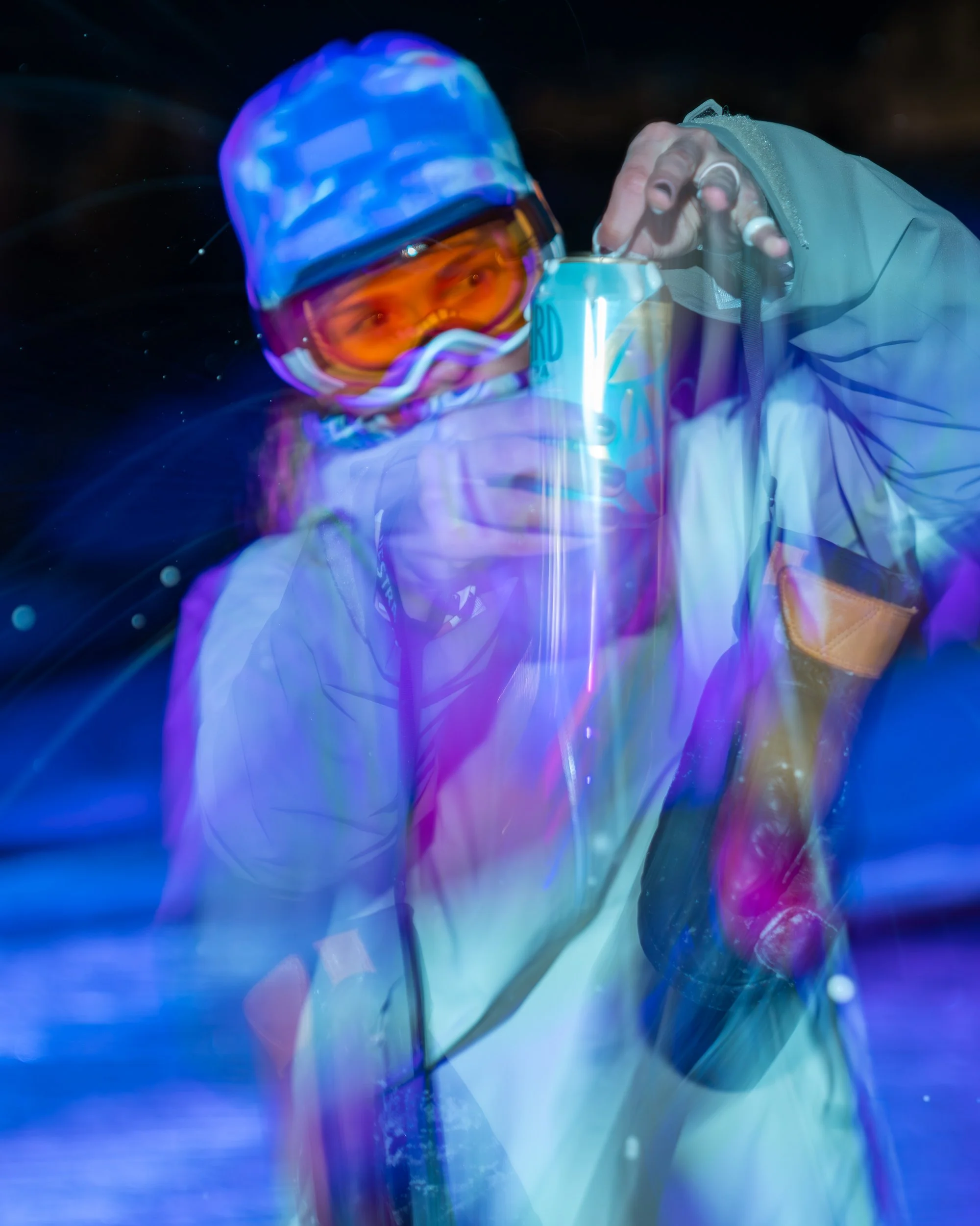 Person wearing a ski jacket, goggles, and helmet, holding a can of beverage in a snowy environment at night, with colorful light effects.