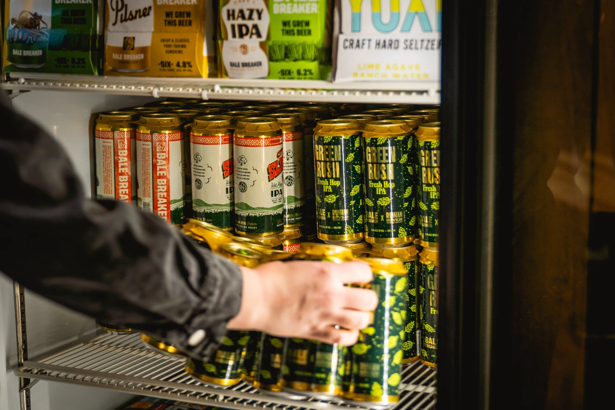 Person reaching for a can of Green Rush Fresh Hop IPA beer in a refrigerator shelf with other canned beers.