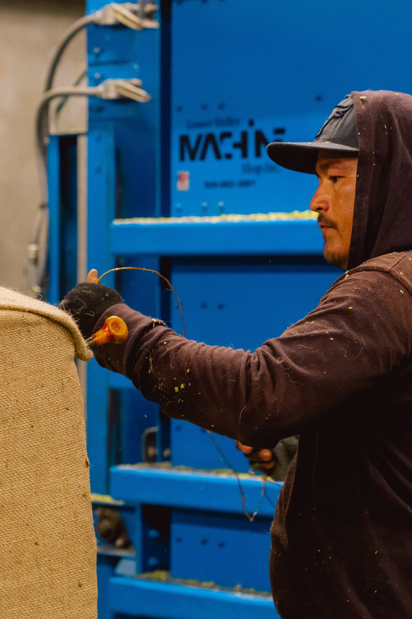 A man wearing a hoodie and gloves is working in a factory or workshop, handling a wire or cord near a burlap sack with a blue industrial machine in the background.