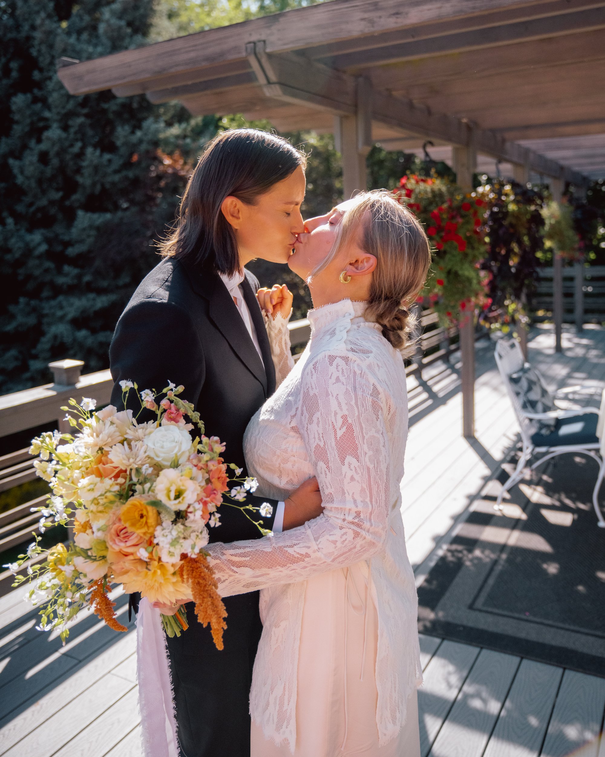 Two women, one in a black suit and the other in a white lace dress, sharing a kiss outdoors, with sunlight on their faces, holding a bouquet of flowers, on a wooden deck with outdoor furniture and greenery in the background.