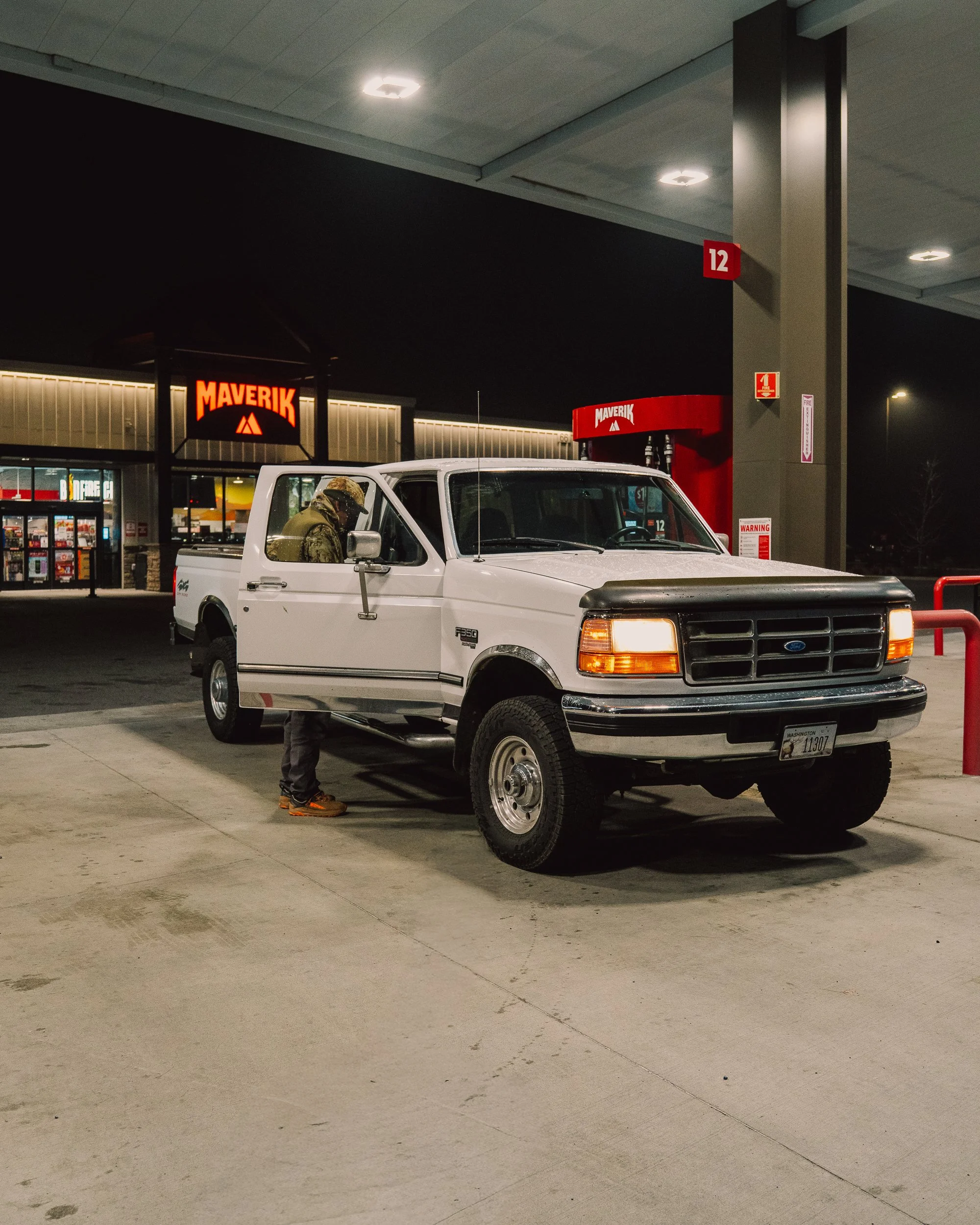 A white Ford pickup truck parked at a gas station at night with a person standing beside it, with the Maverik store visible in the background.