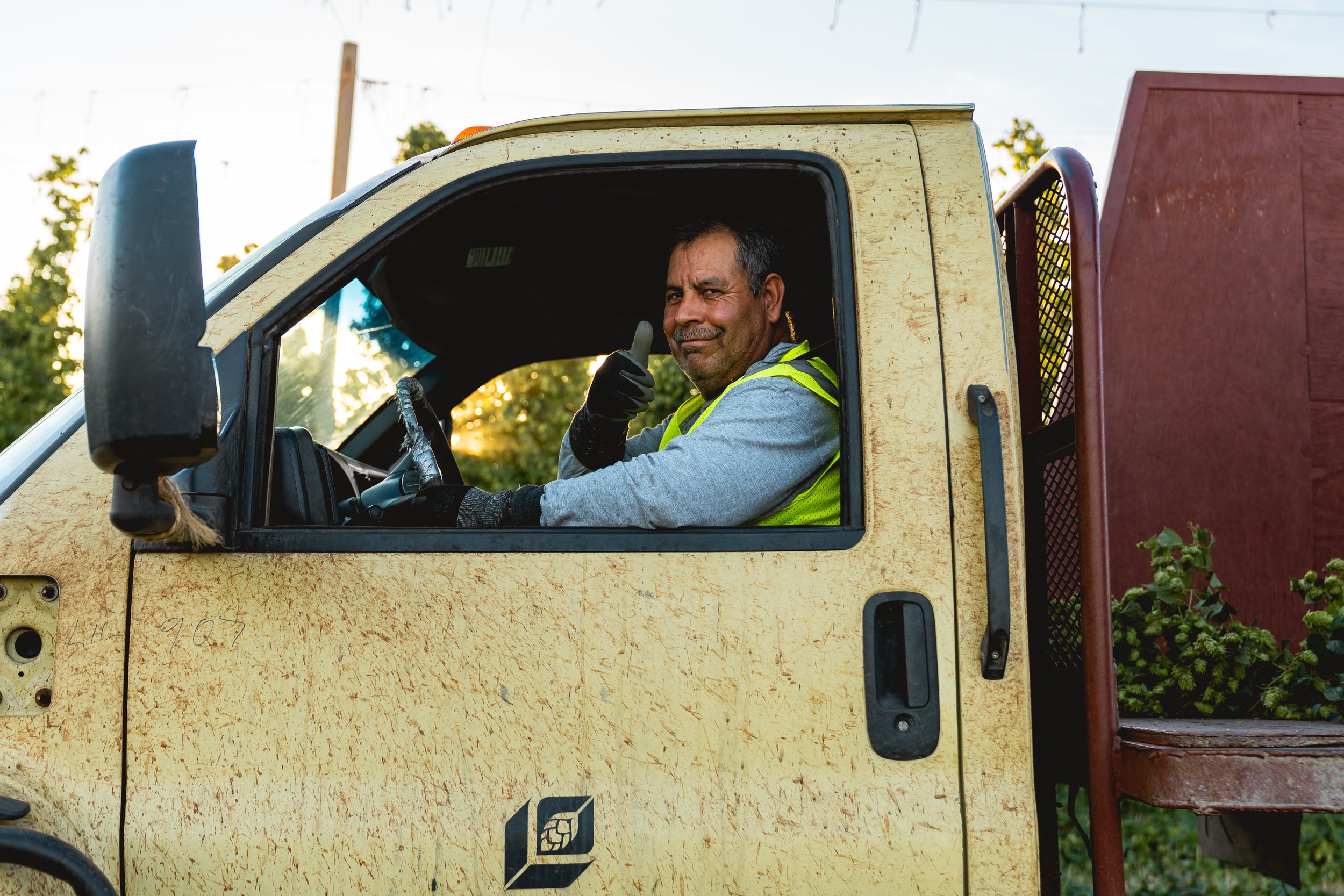 A man in a yellow safety vest sitting inside a yellow work truck, giving a thumbs-up and smiling at the camera.