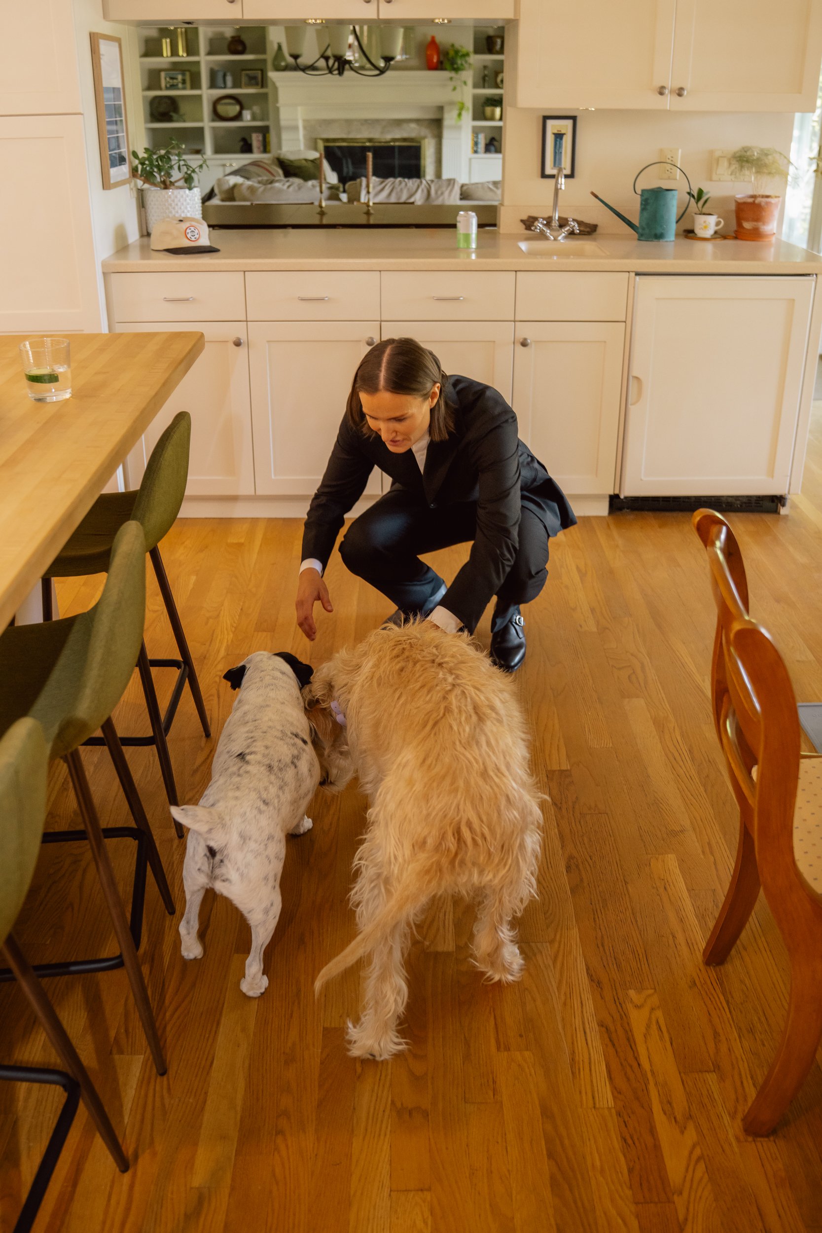 A woman in a black blazer crouches down in a kitchen, feeding two dogs, one white with black spots and the other golden, on hardwood flooring.