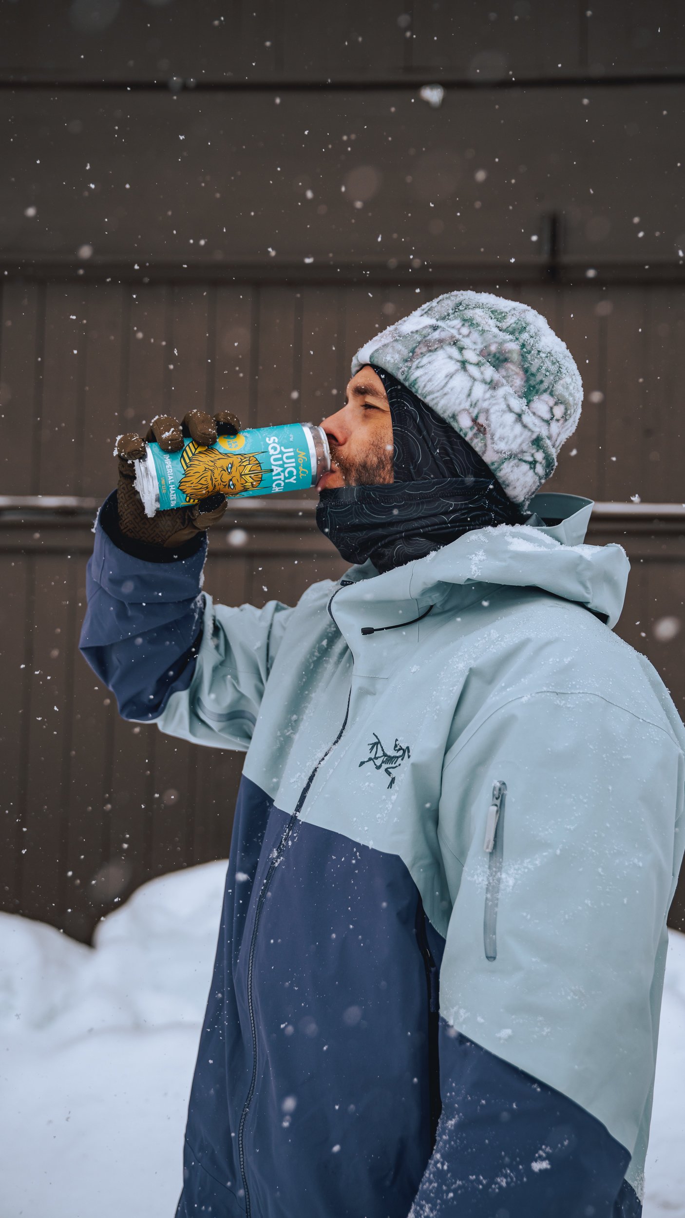 A man in winter clothing drinking from a juice box outdoors in snowy weather.