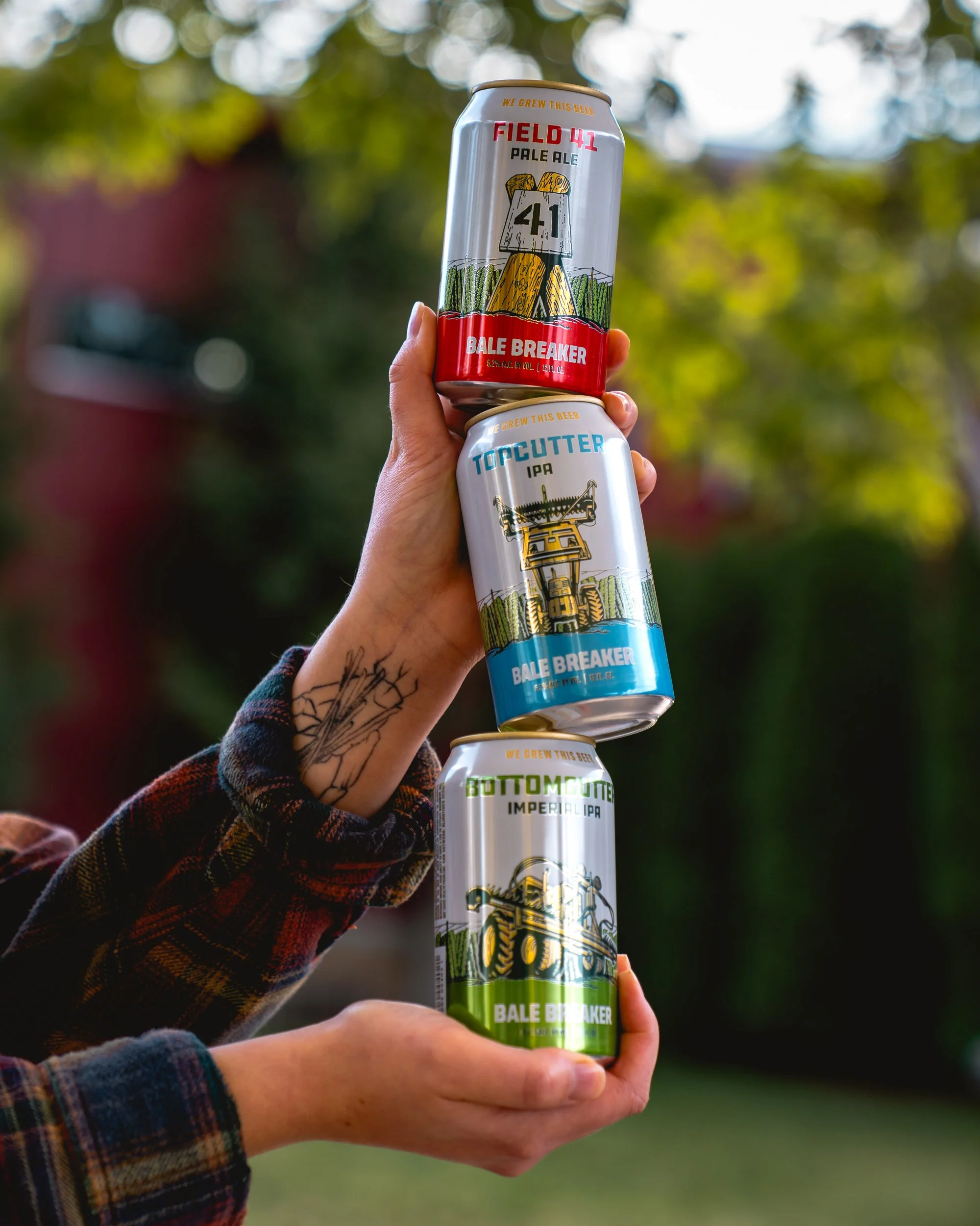 A person holding three cans of Bale Breaker beer, with a background of trees and sky.
