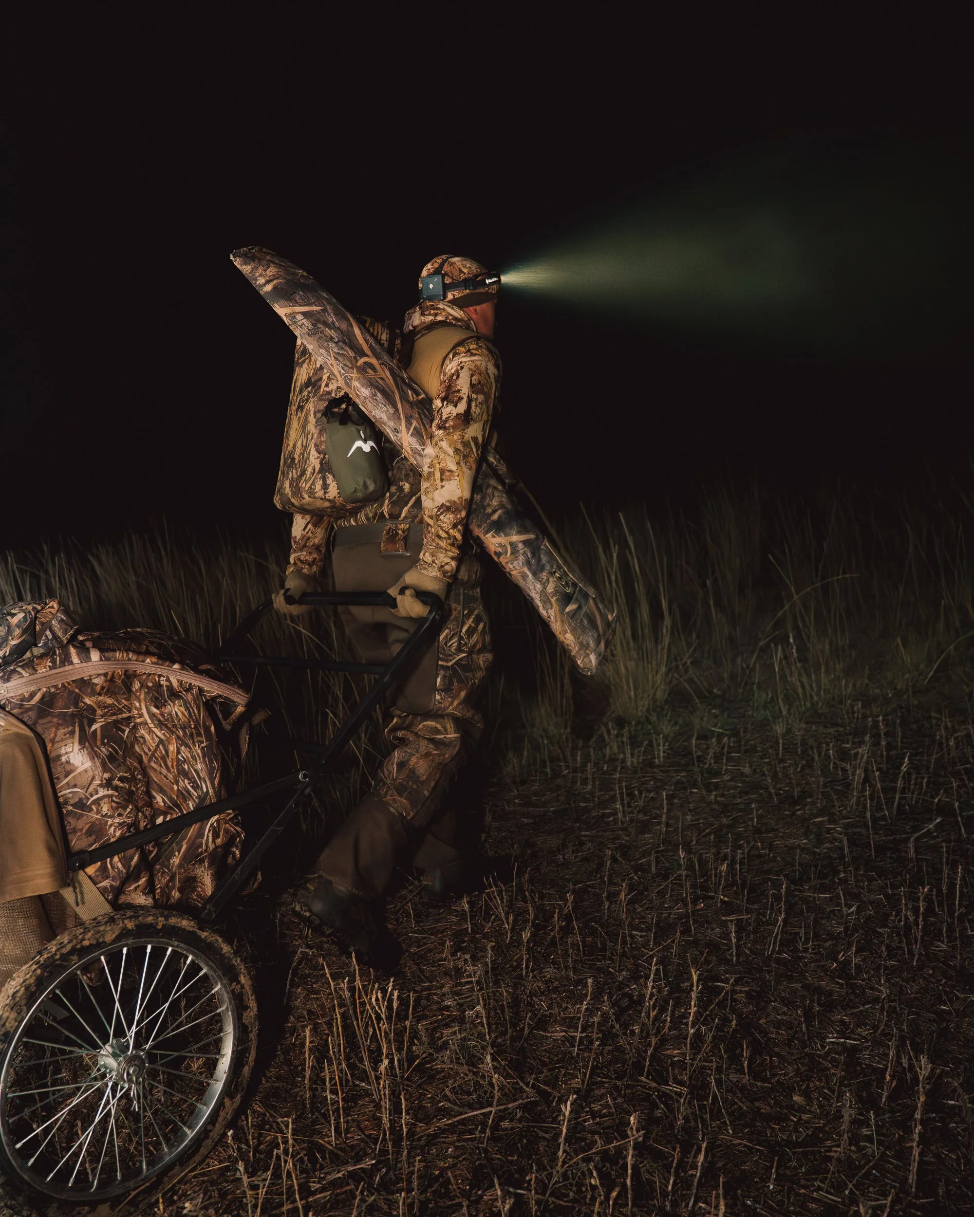 A person dressed in camouflage gear using a night vision headlamp while walking through a field at night, pulling a camouflage backpack on a wheeled cart.