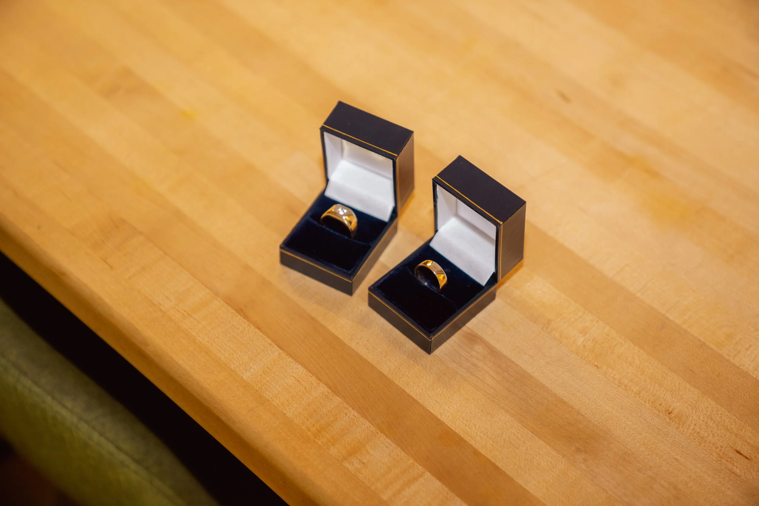 Two black jewelry boxes with gold trim, each containing a gold ring, placed on a wooden surface.