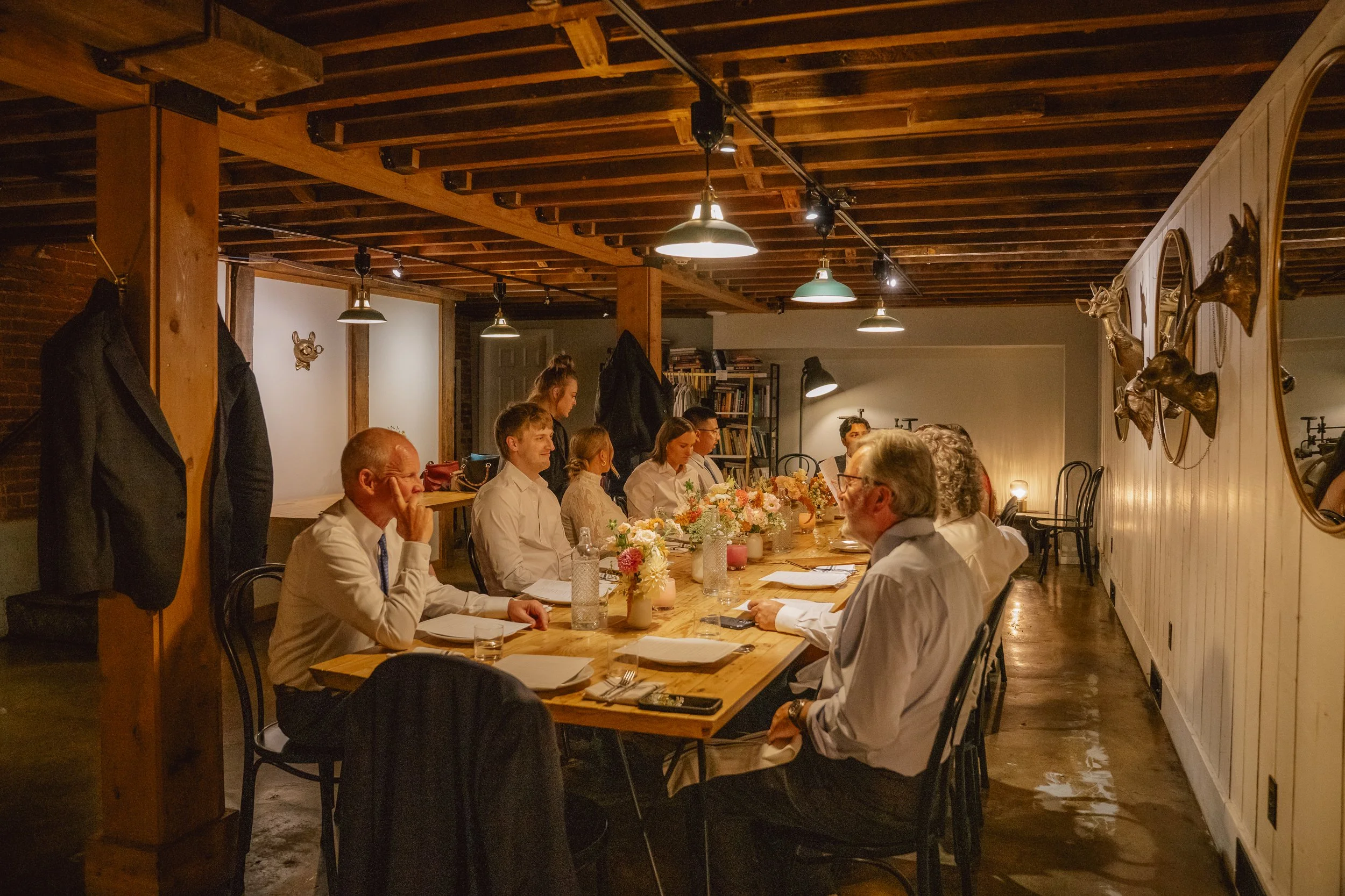A group of people sitting around a long wooden table in a rustic room decorated with mounted animal heads and floral centerpieces, engaging in a meeting or dinner.