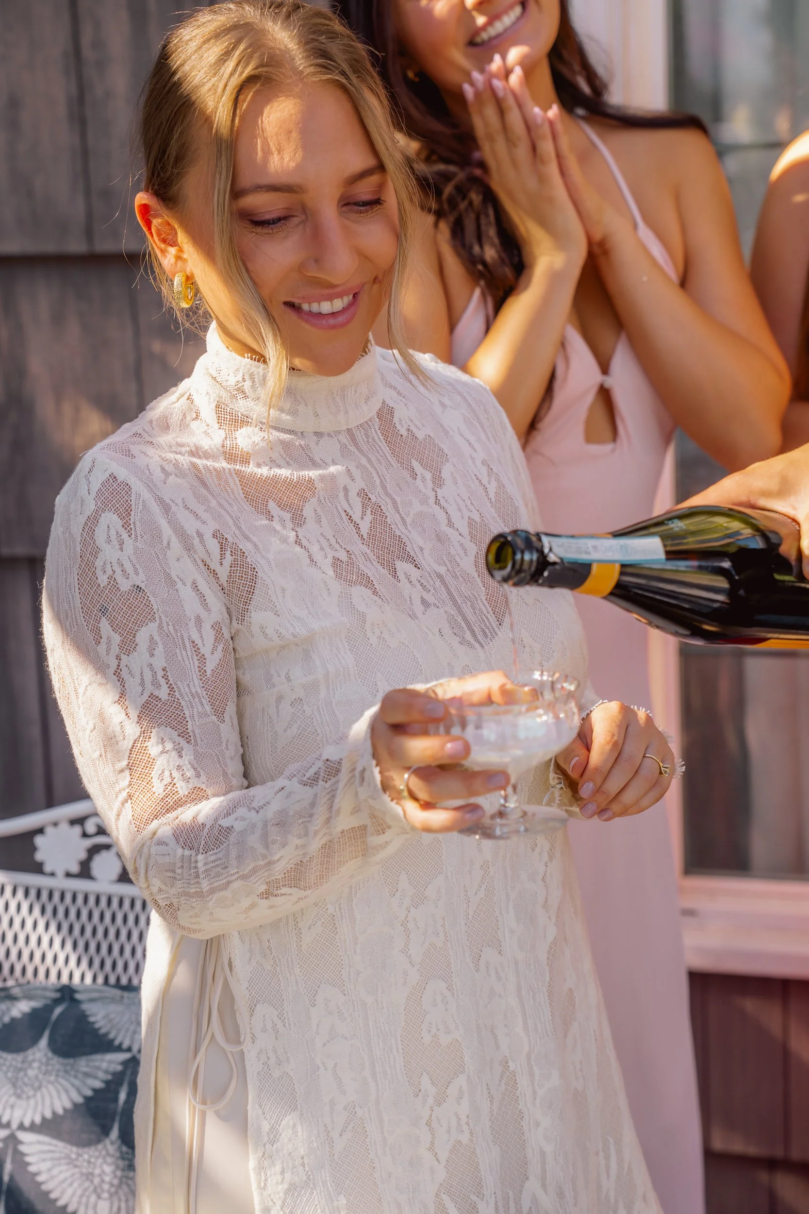 A woman in a lace dress pours champagne into a glass, smiling, while a woman in a pink dress watches and smiles in the background.