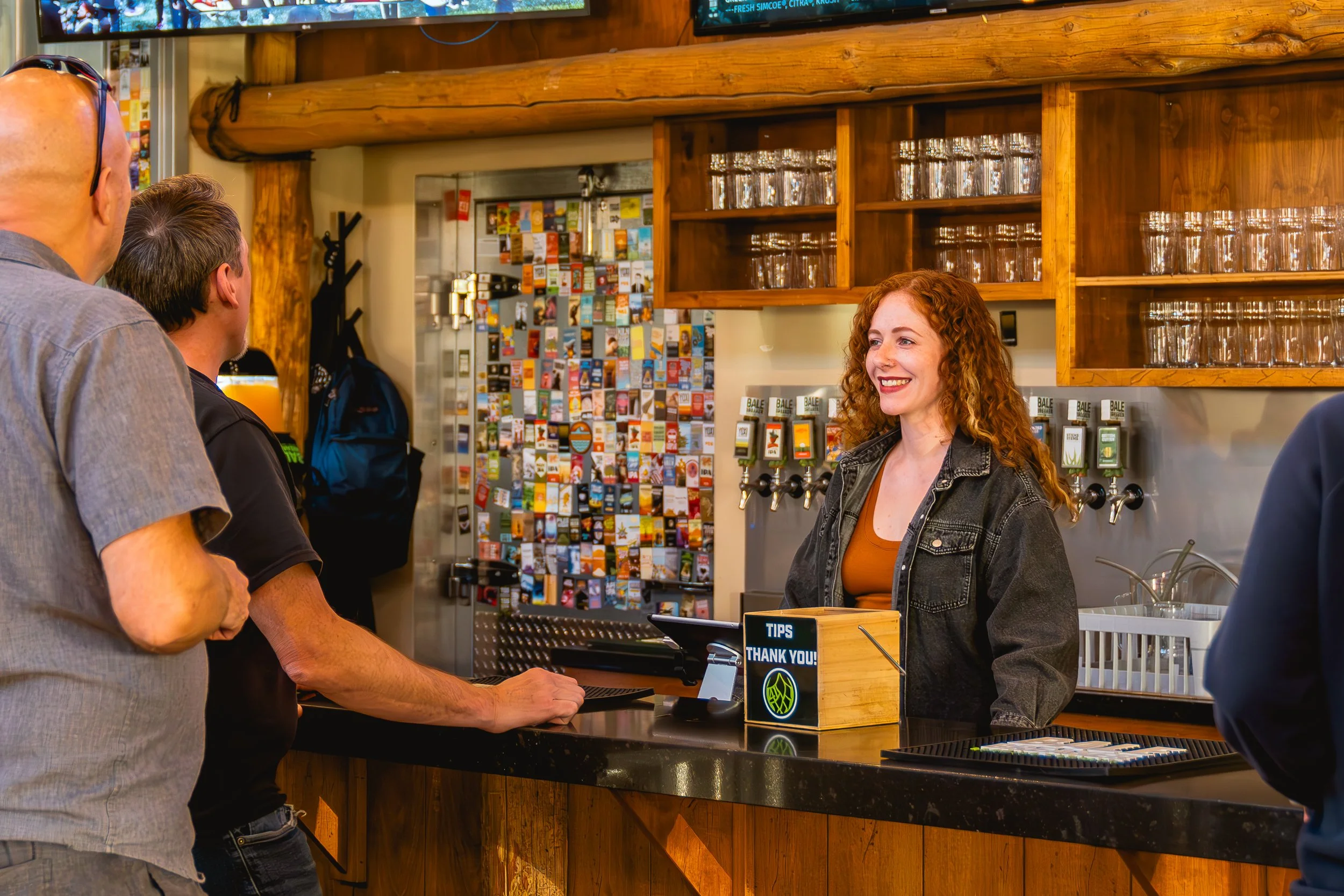 A woman with curly red hair smiling at customers at a bar or restaurant counter, with customers waiting in line and a wooden bar area in the background.
