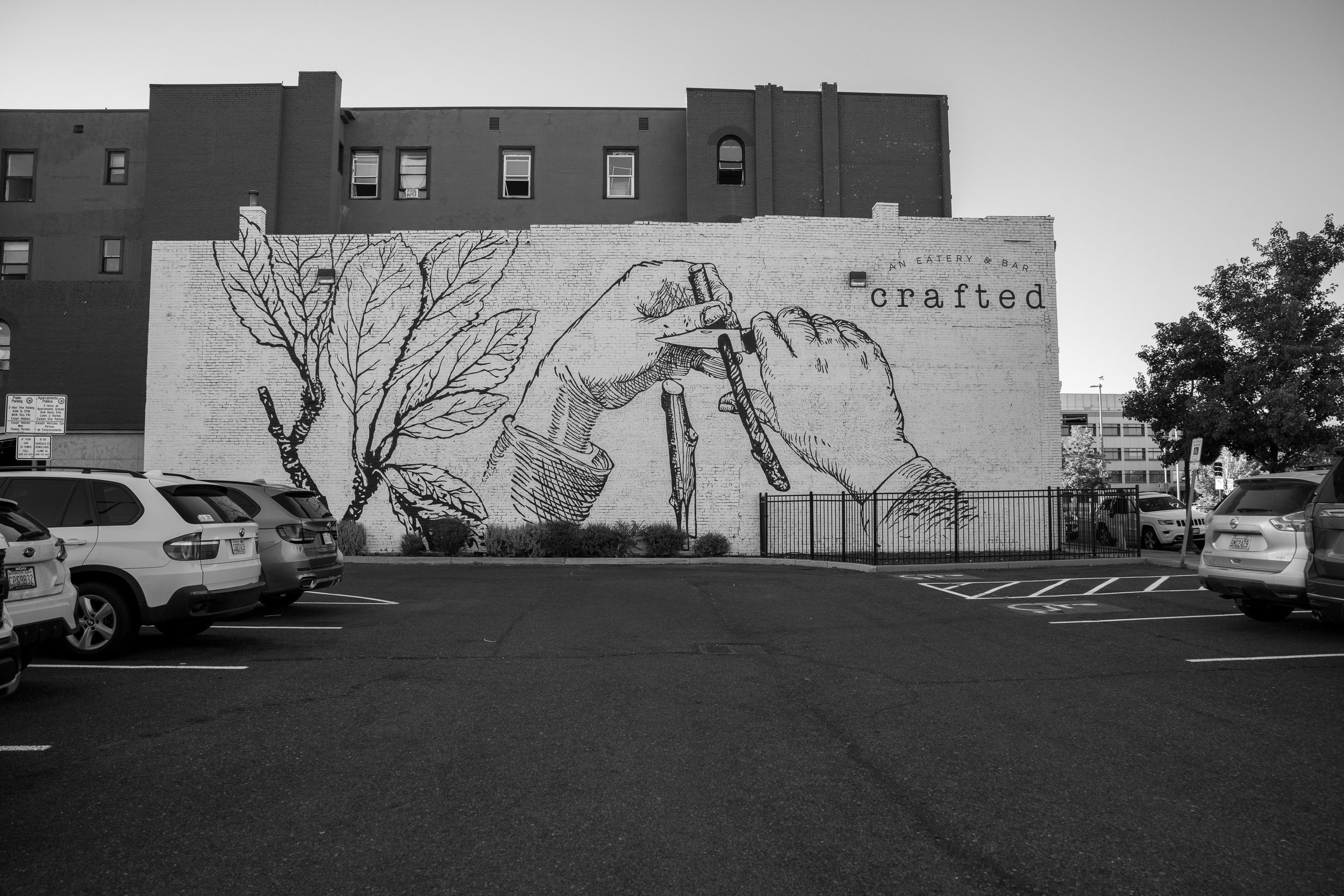 Black and white mural of hands drawing a tree on a brick wall in an urban parking lot.