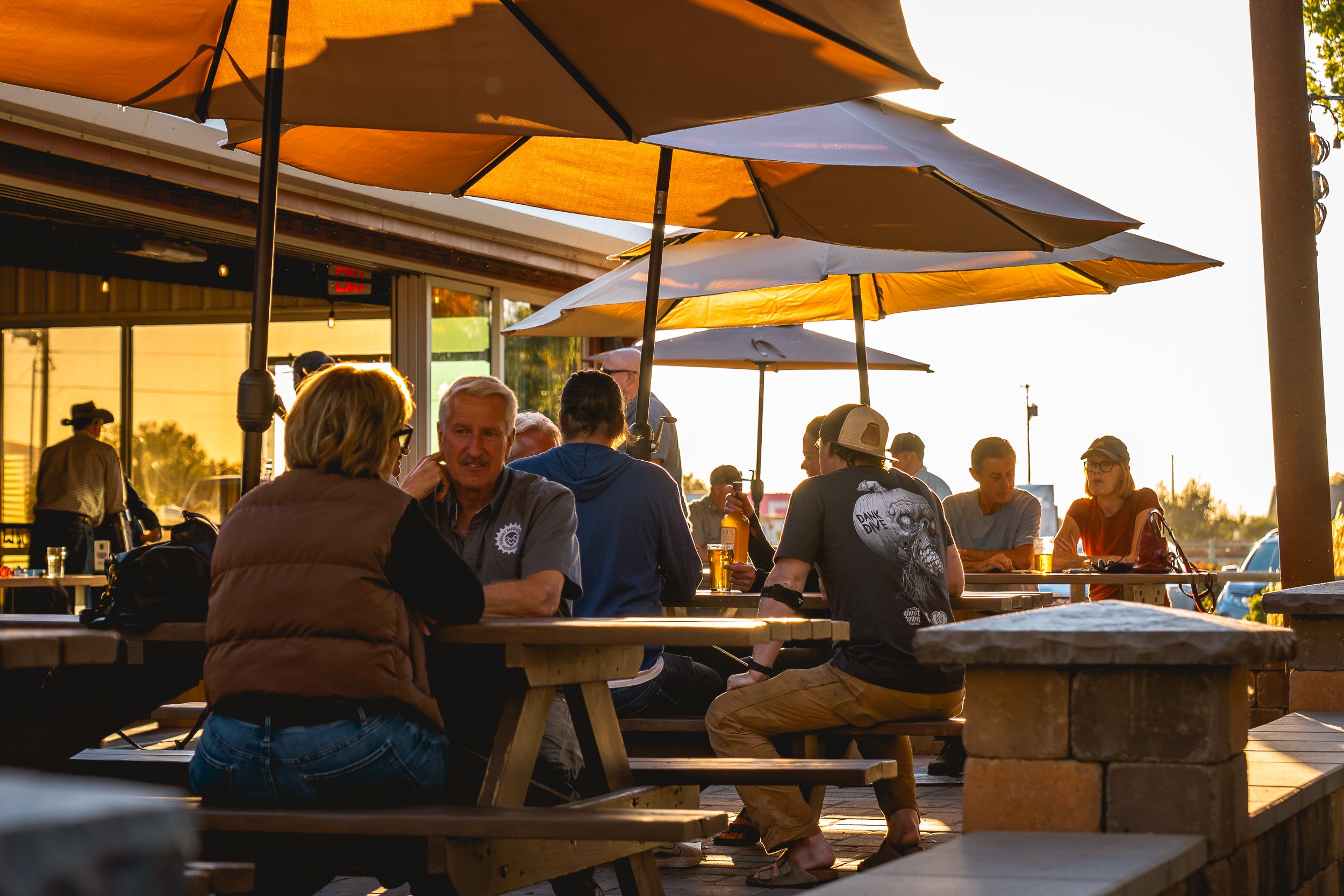 People sitting at outdoor picnic tables under umbrellas at sunset, socializing and enjoying drinks outside a restaurant or bar.