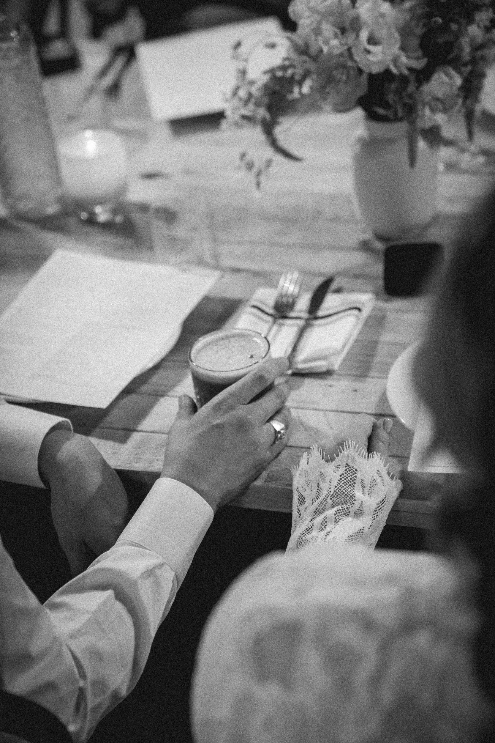 A person in a white shirt holding a glass of beer at a wooden table set with silverware, a menu, a lace napkin, and a bouquet of flowers in a vase.