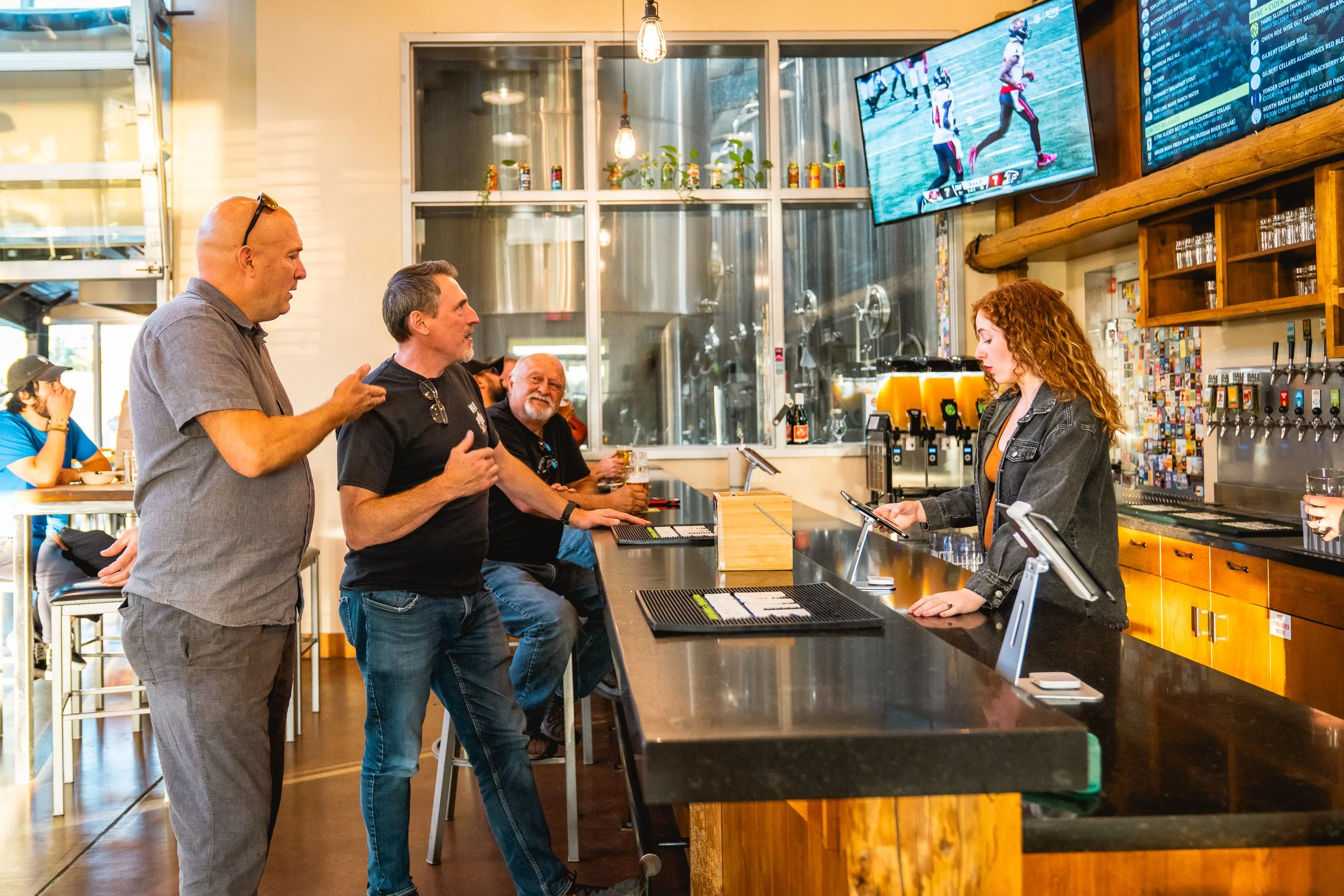 People standing at a bar counter with a bartender. Two men are talking to each other while a third man sits and observes. The bartender is a woman with curly hair, focused on her tablet. There are beer taps, a large screen TV displaying a sports game