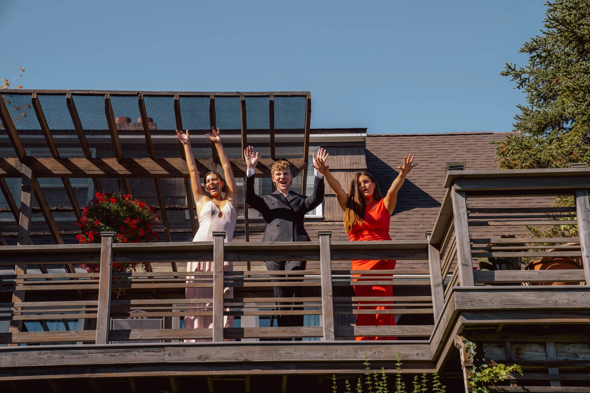 Three people celebrating on a balcony, with two women in dresses and a man in a suit, raising their hands in joy, under a blue sky.