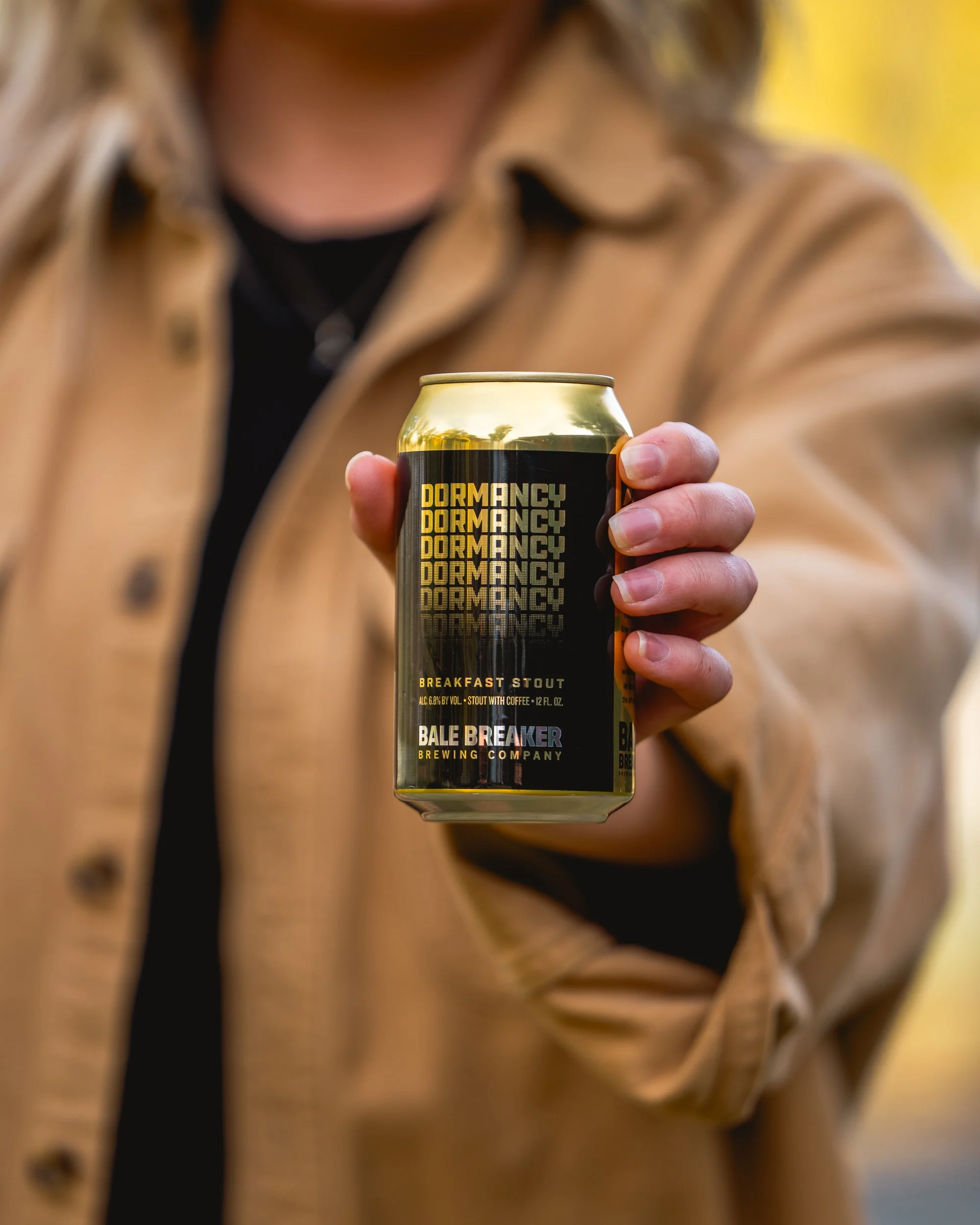 A person in a beige jacket holding a can of Brewdog's Dormancy Breakfast Stout in front of them, with the label prominently displayed.