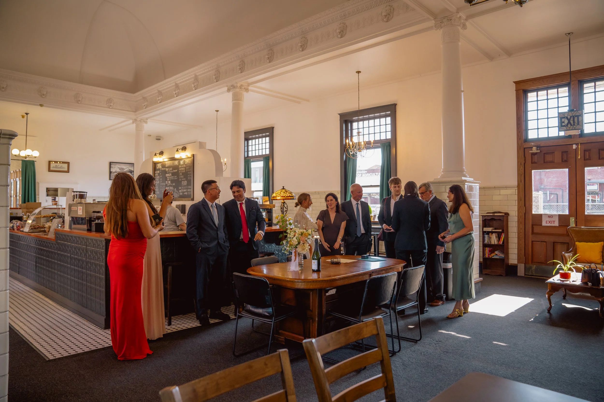 People gathered inside a vintage-style cafe or restaurant with high ceilings, large windows, and classical decor, engaging in conversation, with a person taking photos of others.