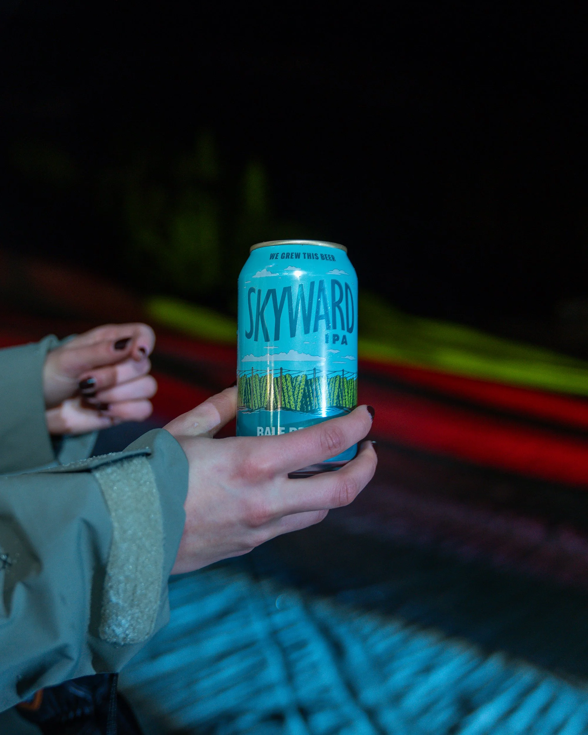 Person holding a can of Skyward IPA beer outdoors at night, with colorful light streaks in the background.