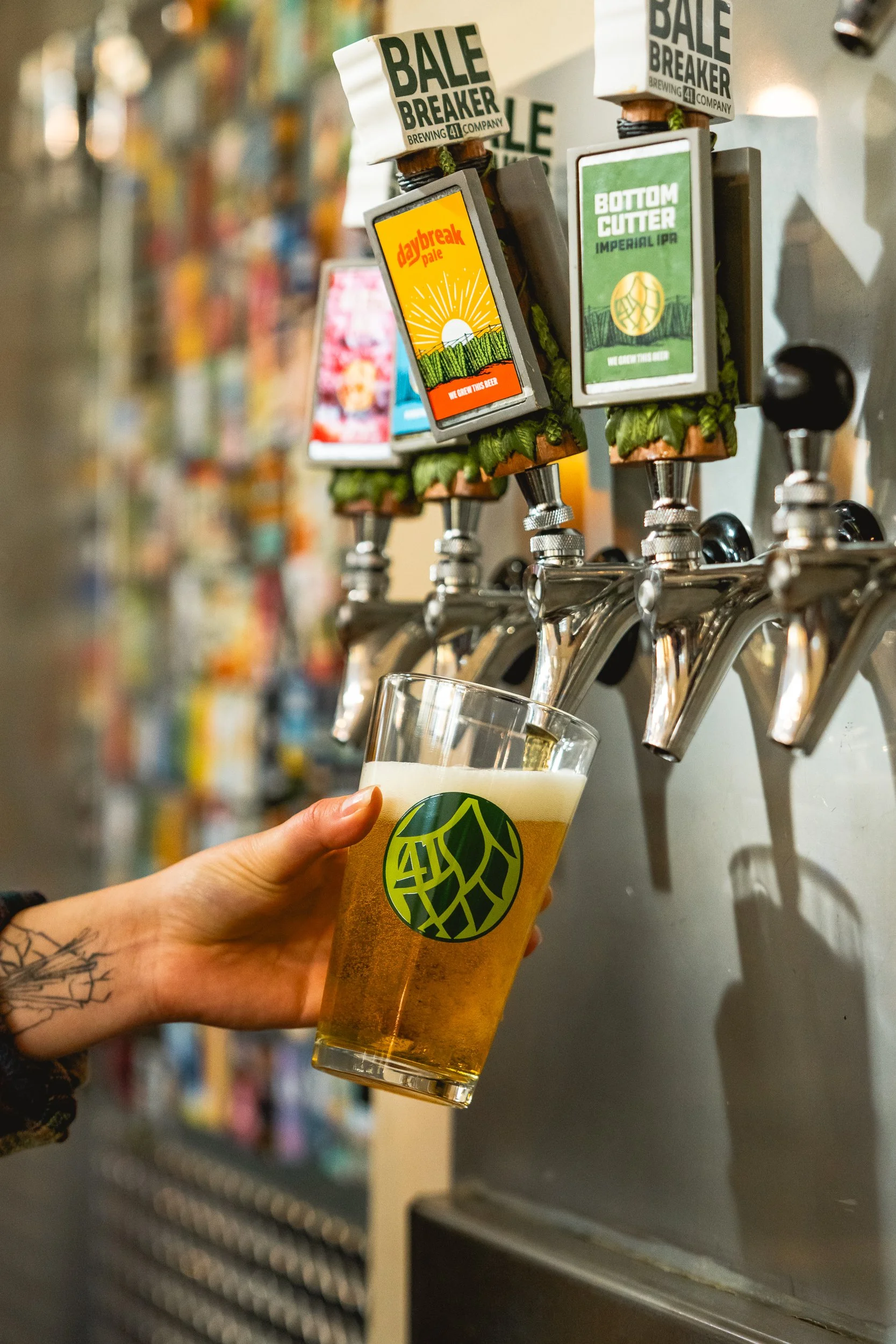A person is filling a plastic cup with beer from a tap at a brewery. The cup has a green logo. Multiple beer taps with digital screens and labels are mounted on a wall, with a blurred background of a store or brewery interior.