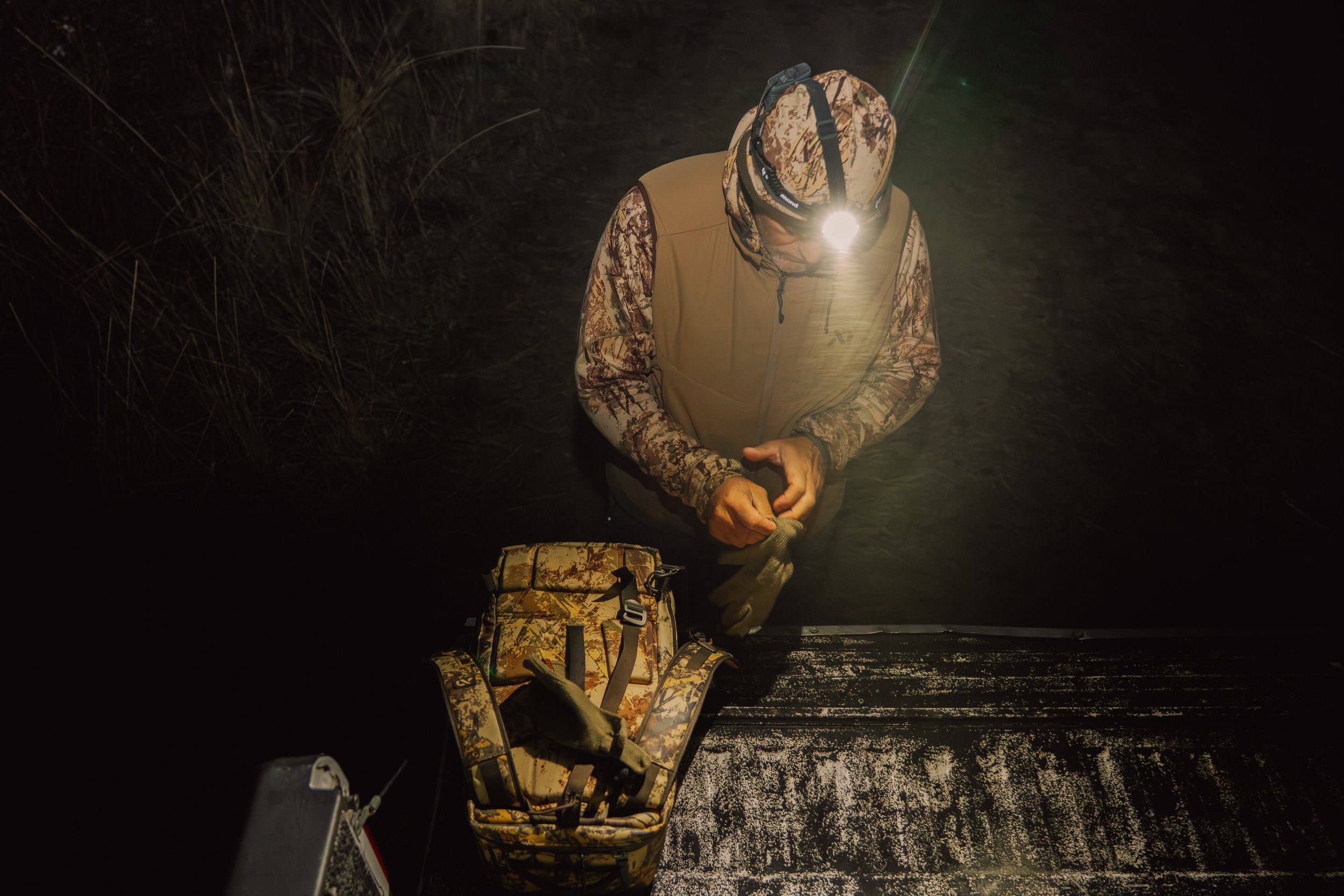 A person wearing camouflage patterned clothing, a vest, and a headlamp is standing in shallow water at night, holding a fishing line. There is a camouflage backpack on the surface near them.