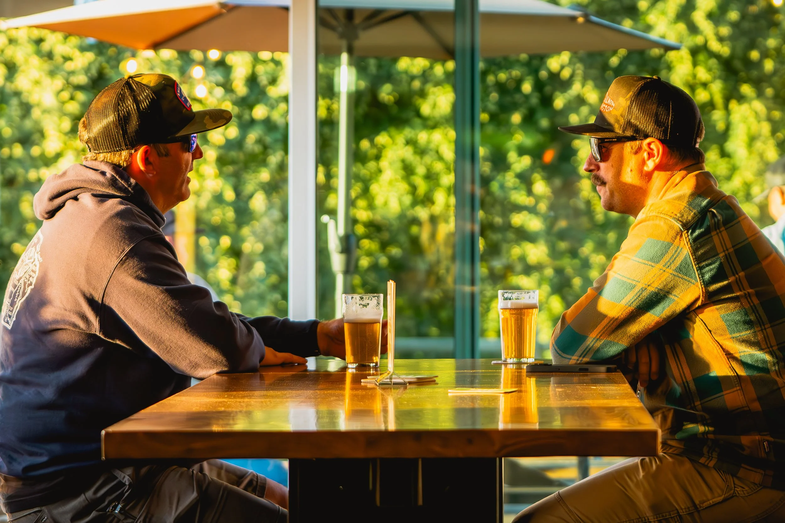 Two men sitting at a table with glasses of beer in an outdoor setting, engaging in conversation. They are wearing casual clothing and caps, with sunlight and greenery in the background.