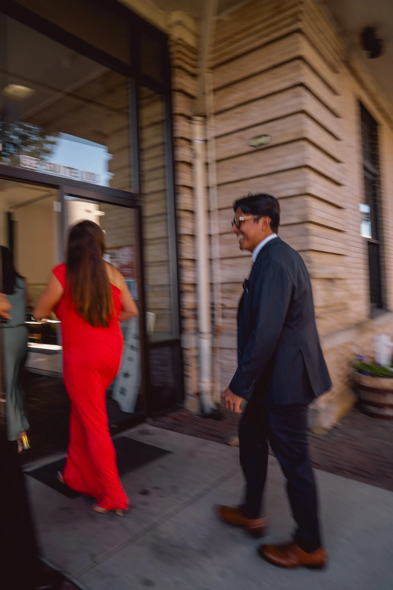 A man in a dark suit with sunglasses, smiling, walking towards the entrance of a building behind a woman wearing a red dress. The building has brick walls and large glass doors.