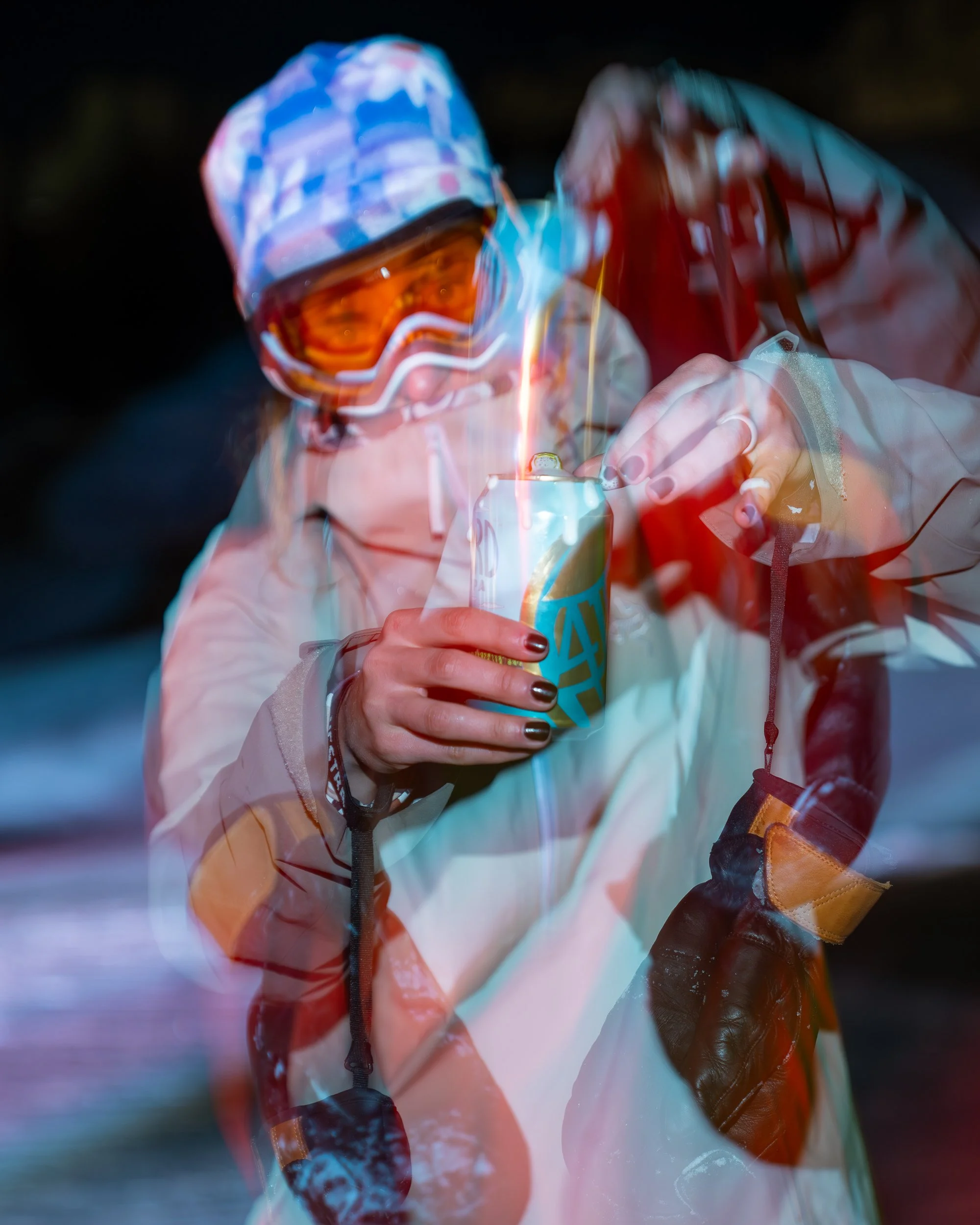 A person in winter gear and ski goggles is holding a can of beer while lighting it with a match, with sparks visible.