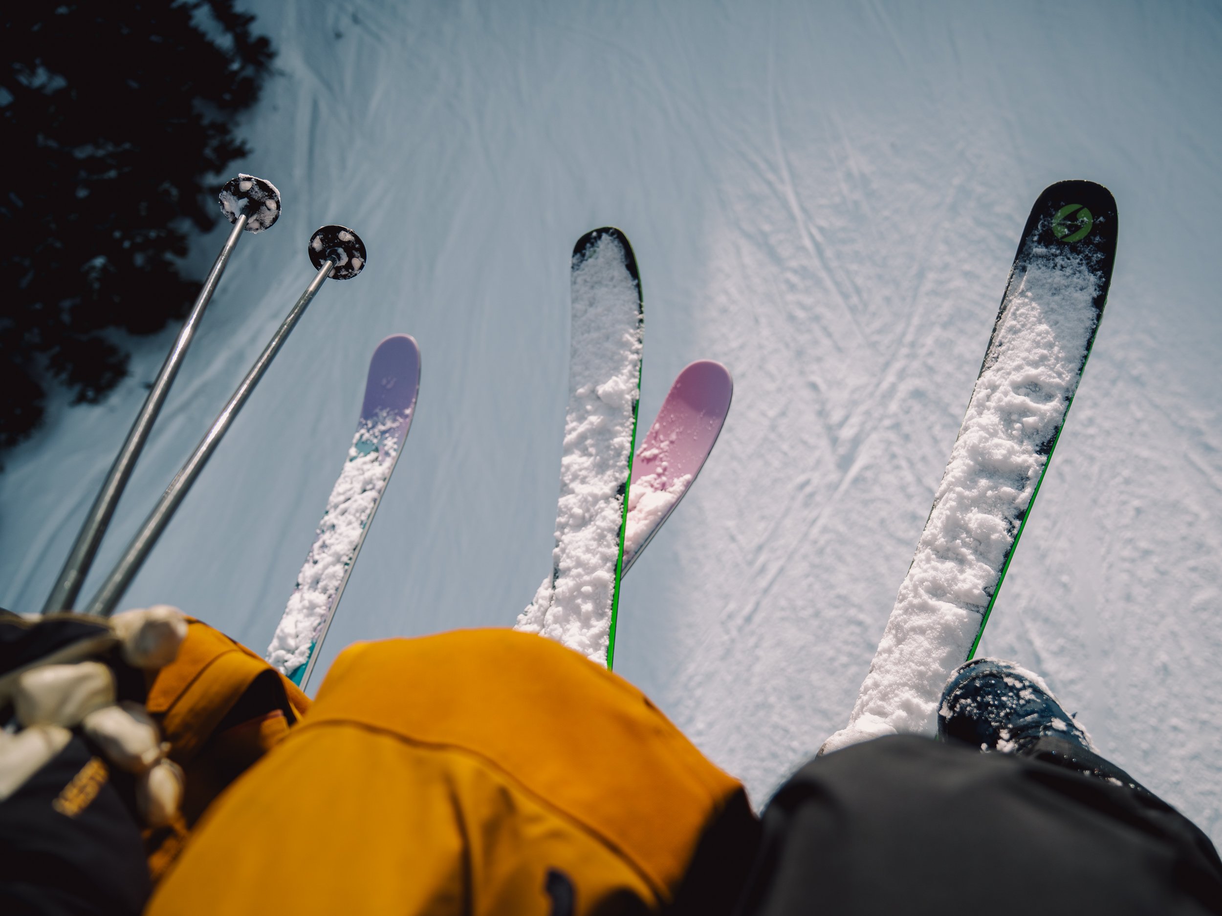 View of skis and ski poles on snow-covered ground, with a person wearing yellow and black clothing standing nearby.