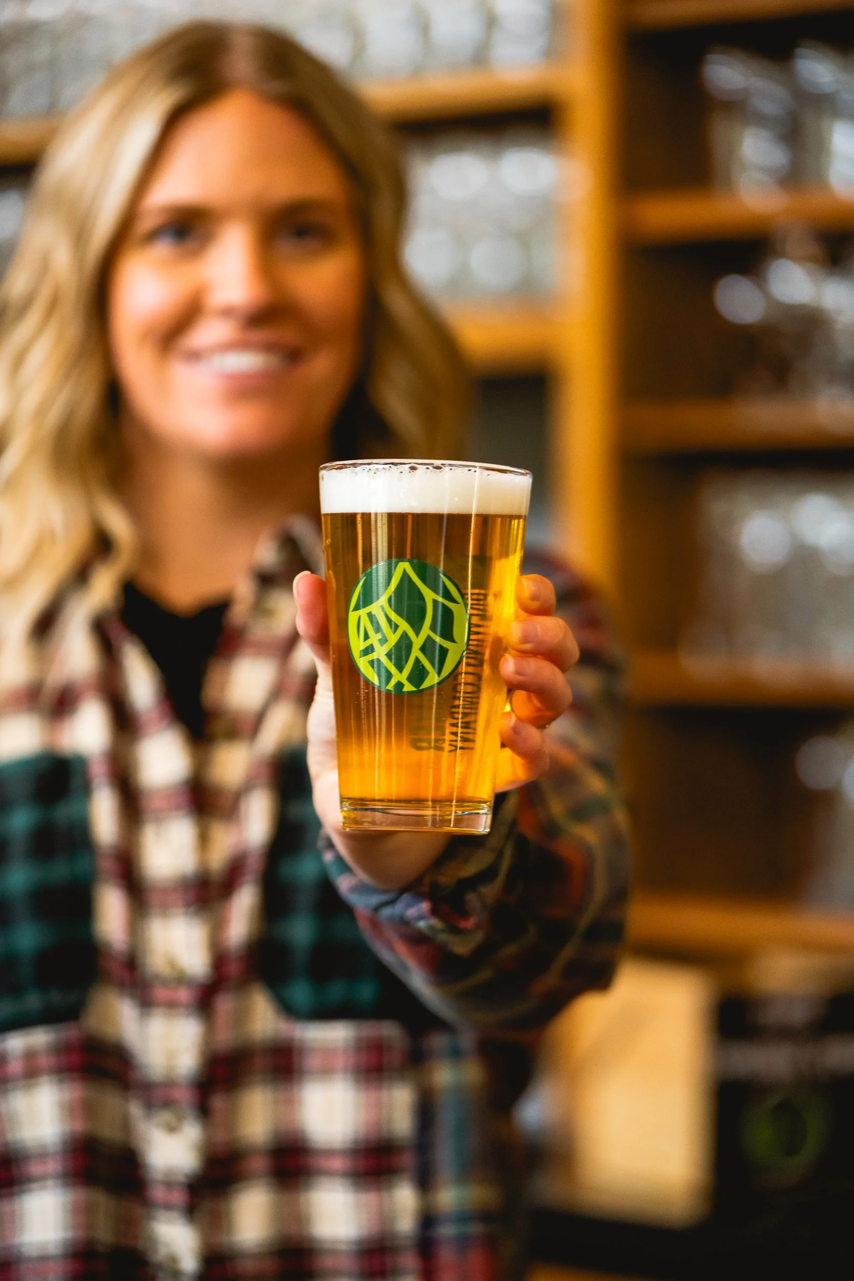 A woman smiling and holding up a glass of beer with a logo on it, in a cozy setting with shelves in the background.