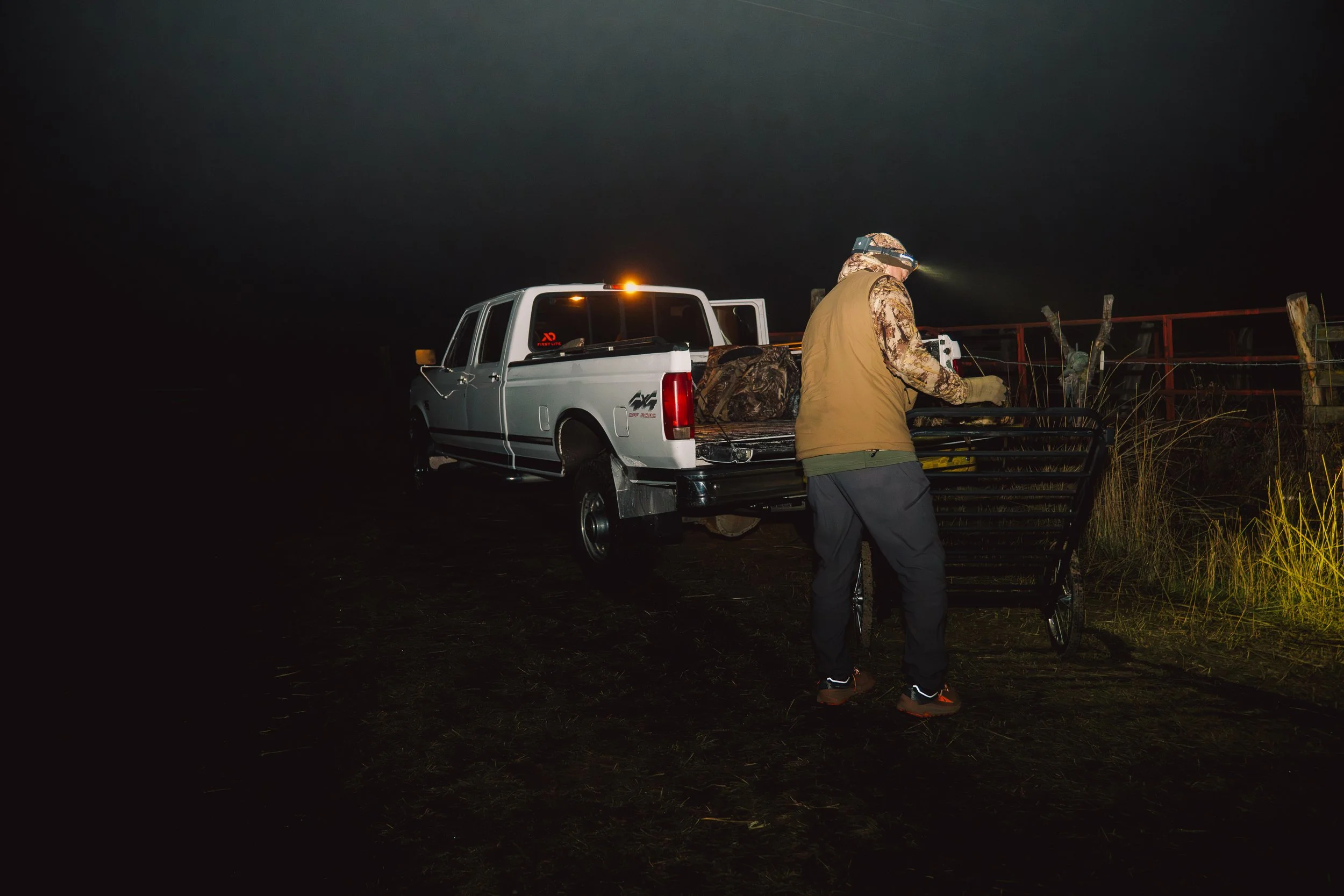 Person with headlamp loading supplies into a pickup truck at night, with darkness surrounding.