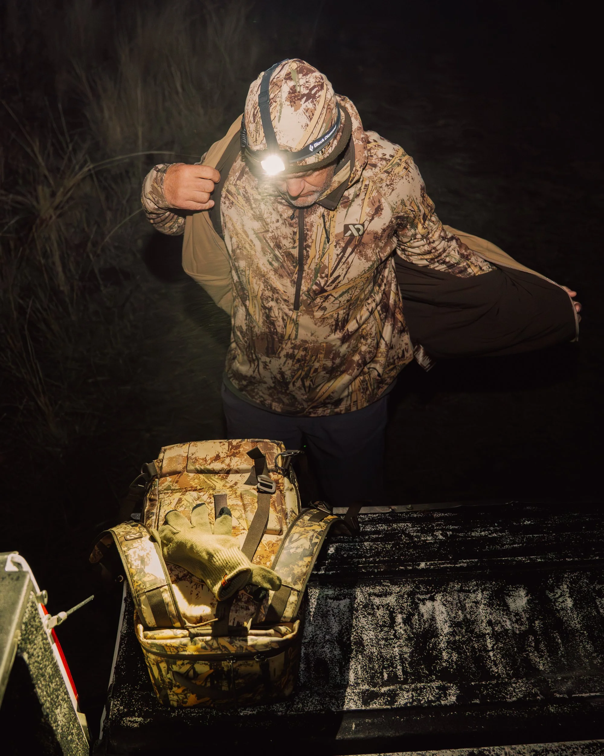 Man wearing camouflage jacket and headlamp standing in water at night, with a camouflage bag and gloves on wooden platform.