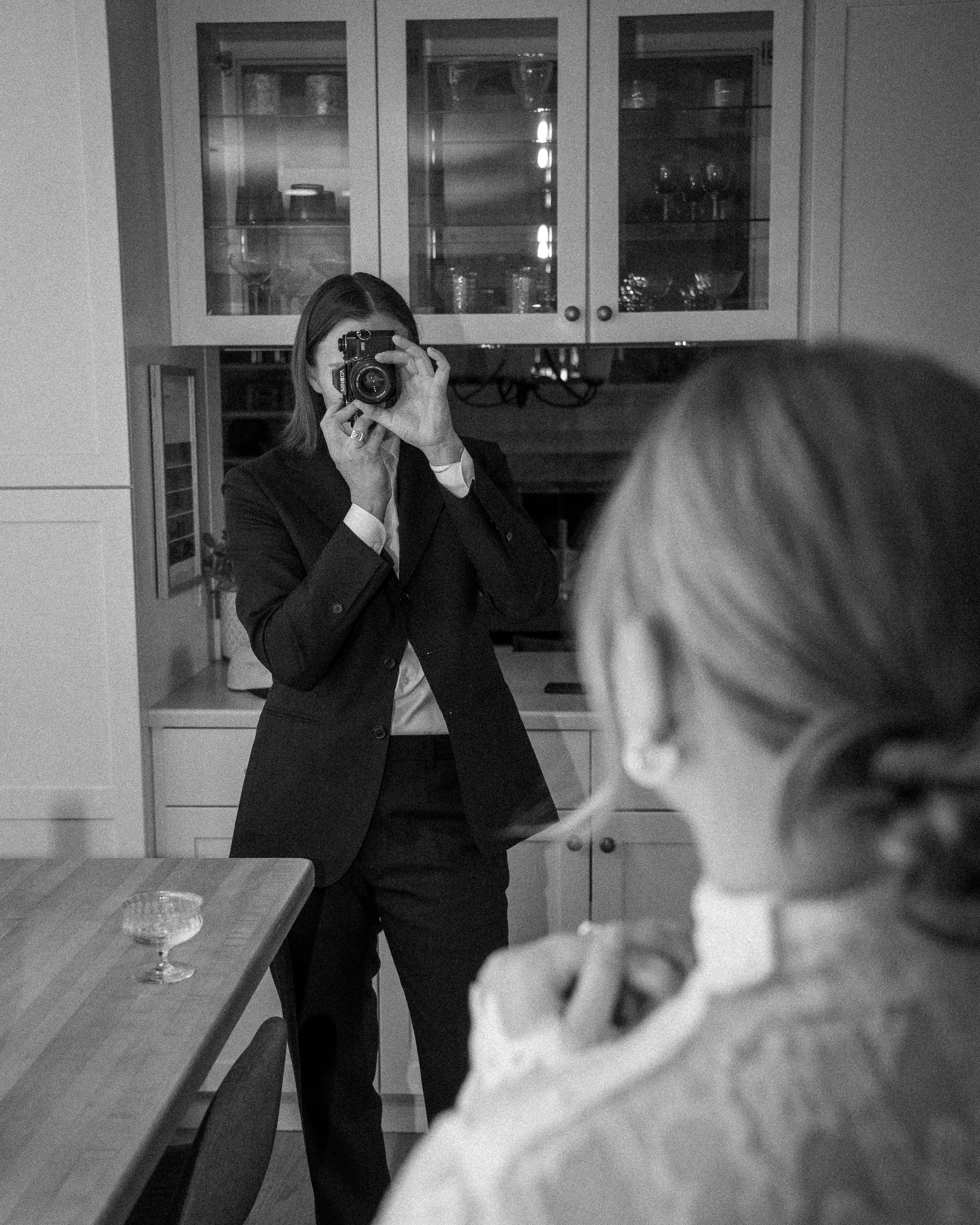 Black and white photo of a woman in a dark suit taking a picture of a woman with glasses in a kitchen.