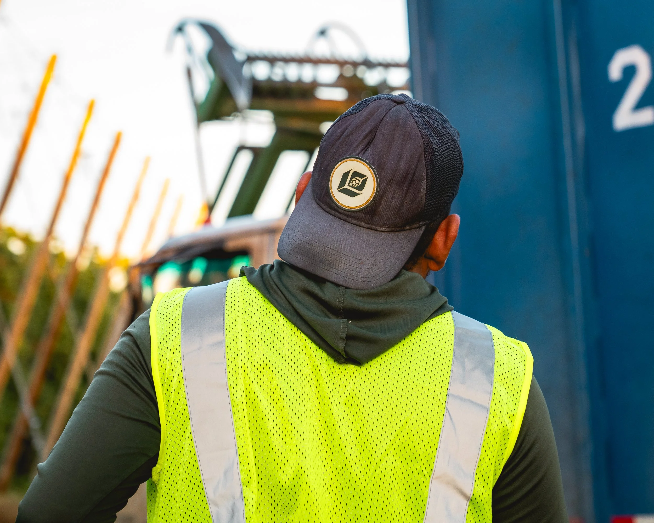 A person wearing a yellow safety vest and a black cap with a logo, facing away outdoors.