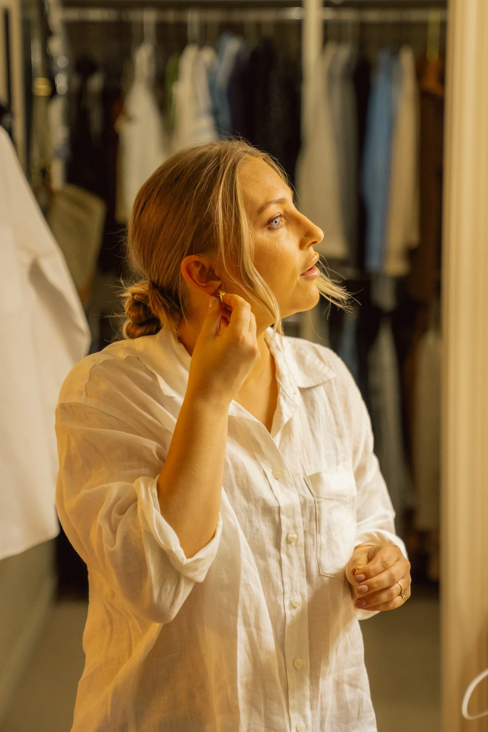 A woman with blonde hair in a loose bun putting on an earring in front of a mirror inside a walk-in closet or dressing room.