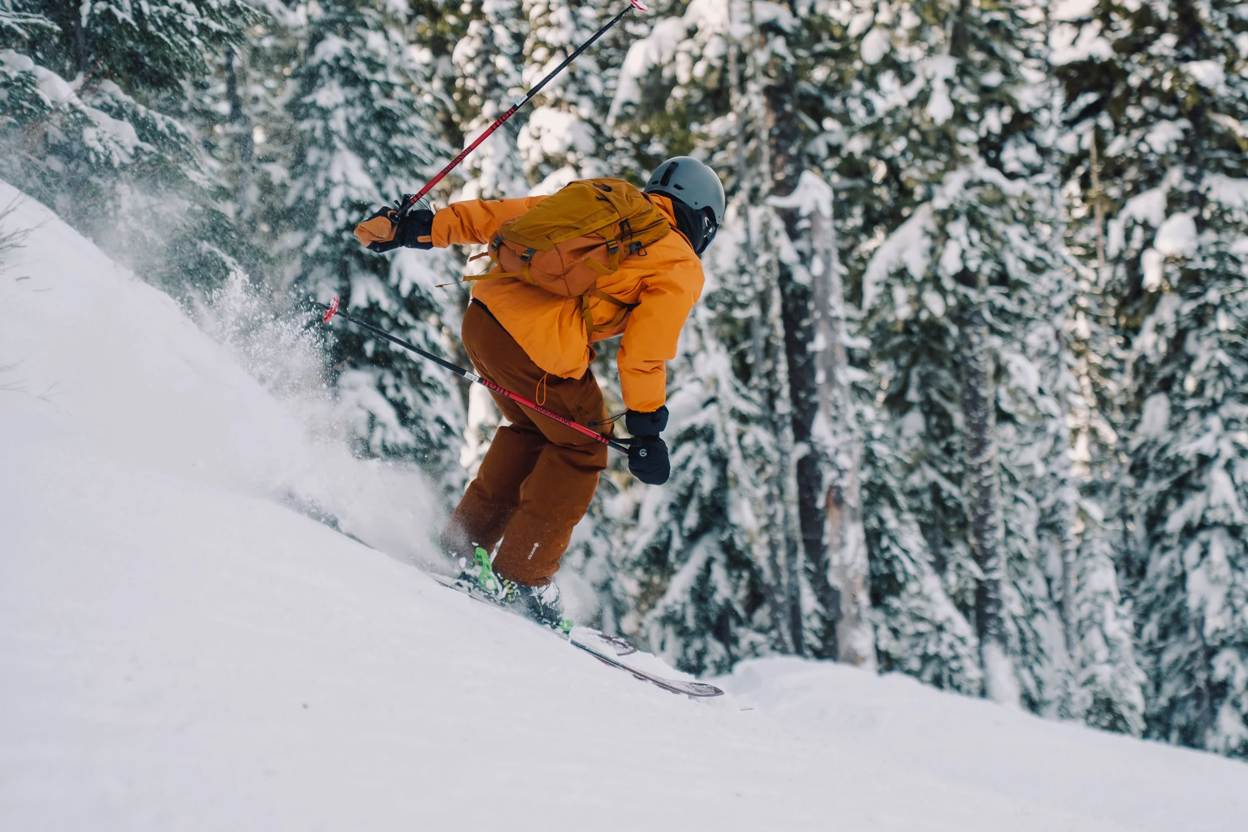 Skier wearing an orange jacket, brown pants, a gray helmet, and a yellow backpack skiing down a snowy slope with snow-covered trees in the background.