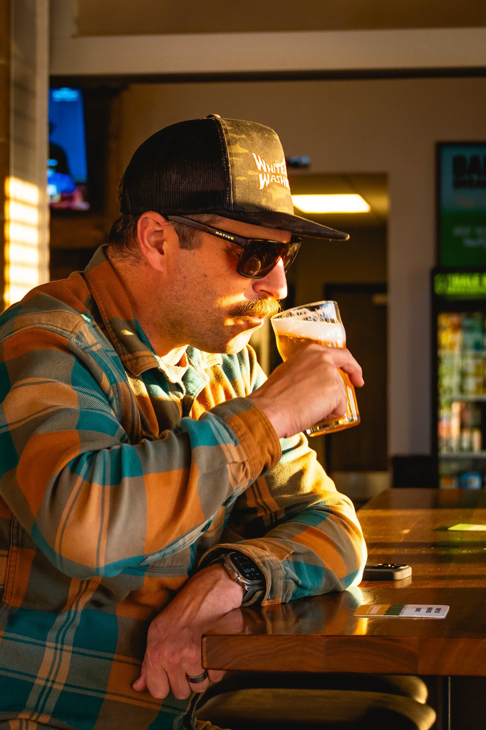A man with a mustache, wearing sunglasses, a checkered shirt, a black cap with camouflage and lettering, and a watch, drinking a beer at a bar or pub during sunset or late afternoon.