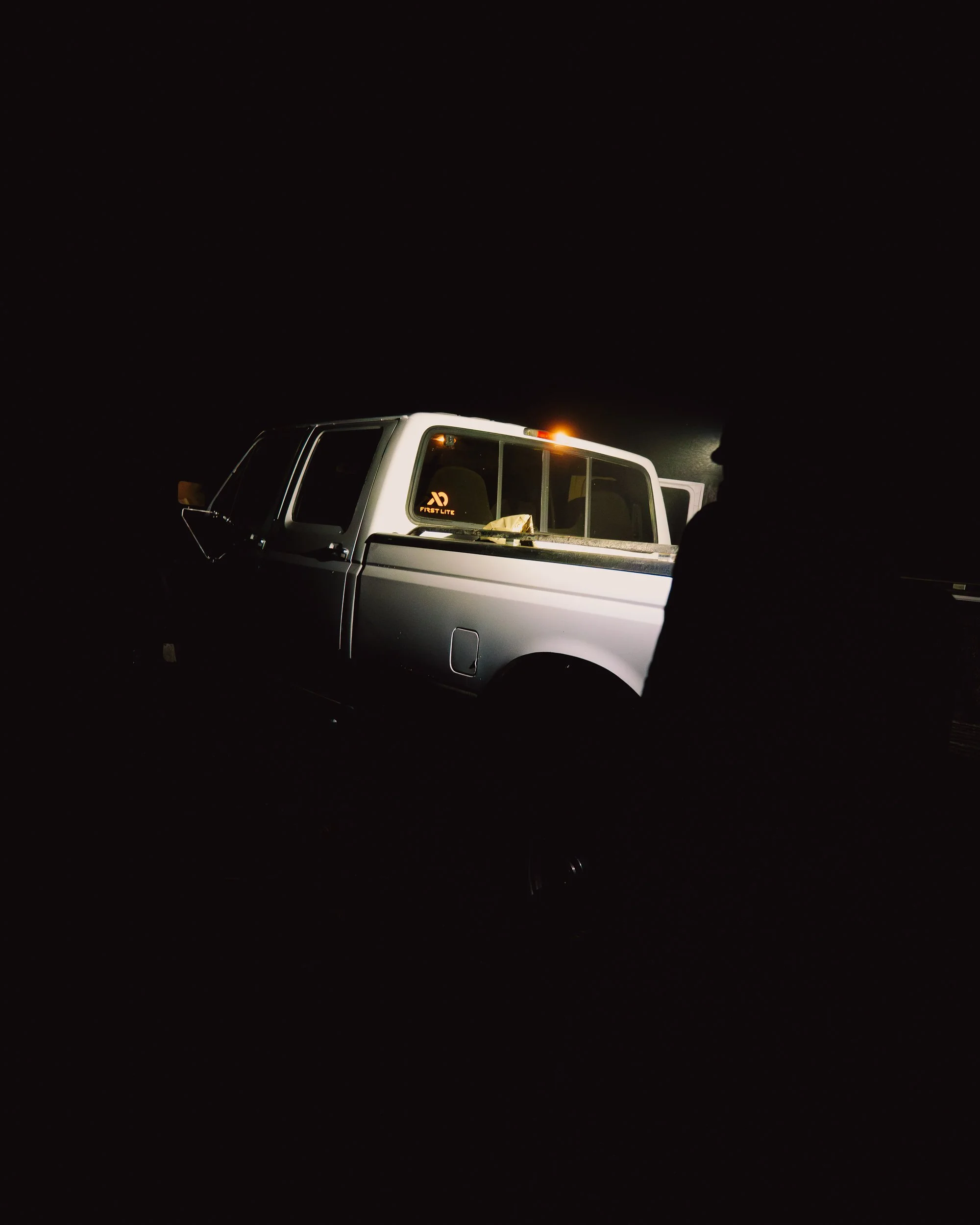 A pickup truck in a dark setting with its front illuminated, showing part of the interior and a silhouette of a person nearby.