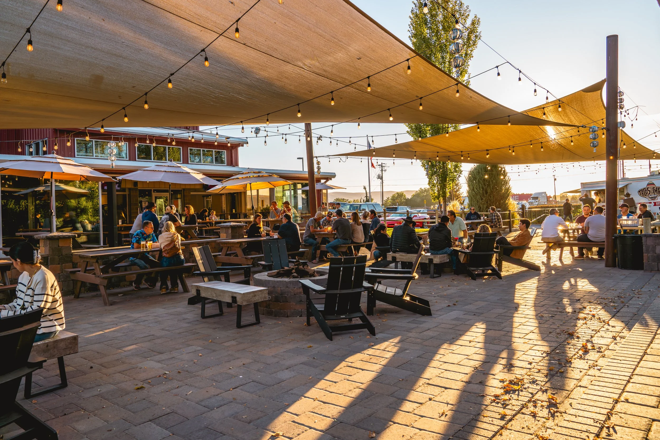 People dining outdoors at a restaurant patio with string lights, umbrellas, and shade sails during sunset.