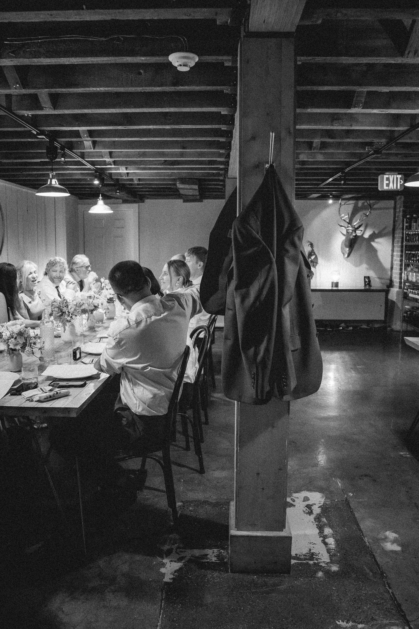 A black-and-white photo of a restaurant or dining area with people sitting at a long table. There are floral arrangements on the table, and a coat hanging on a pillar in the foreground. The ceiling has exposed wooden beams and hanging lights, with an