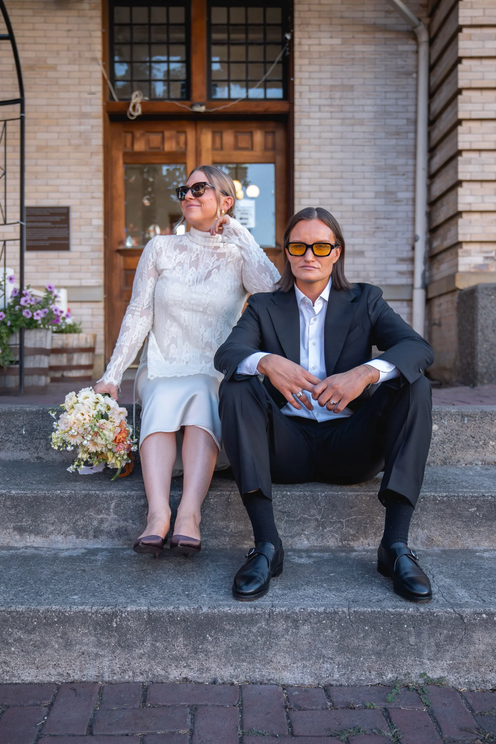 A woman in a white lace blouse and beige skirt holding a bouquet of flowers, and a woman in a black suit and sunglasses sitting on steps outside a brick building.
