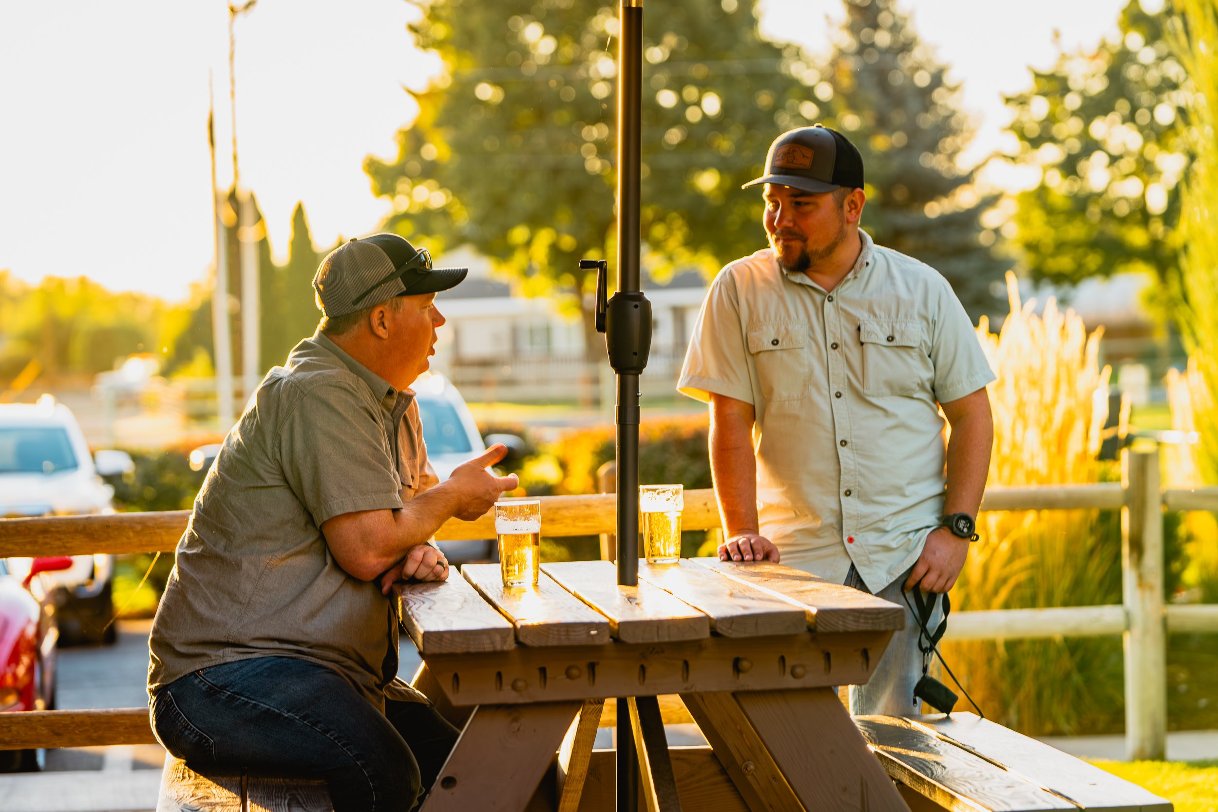 Two men having a conversation outdoors at a picnic table with two glasses of beer, during sunset.