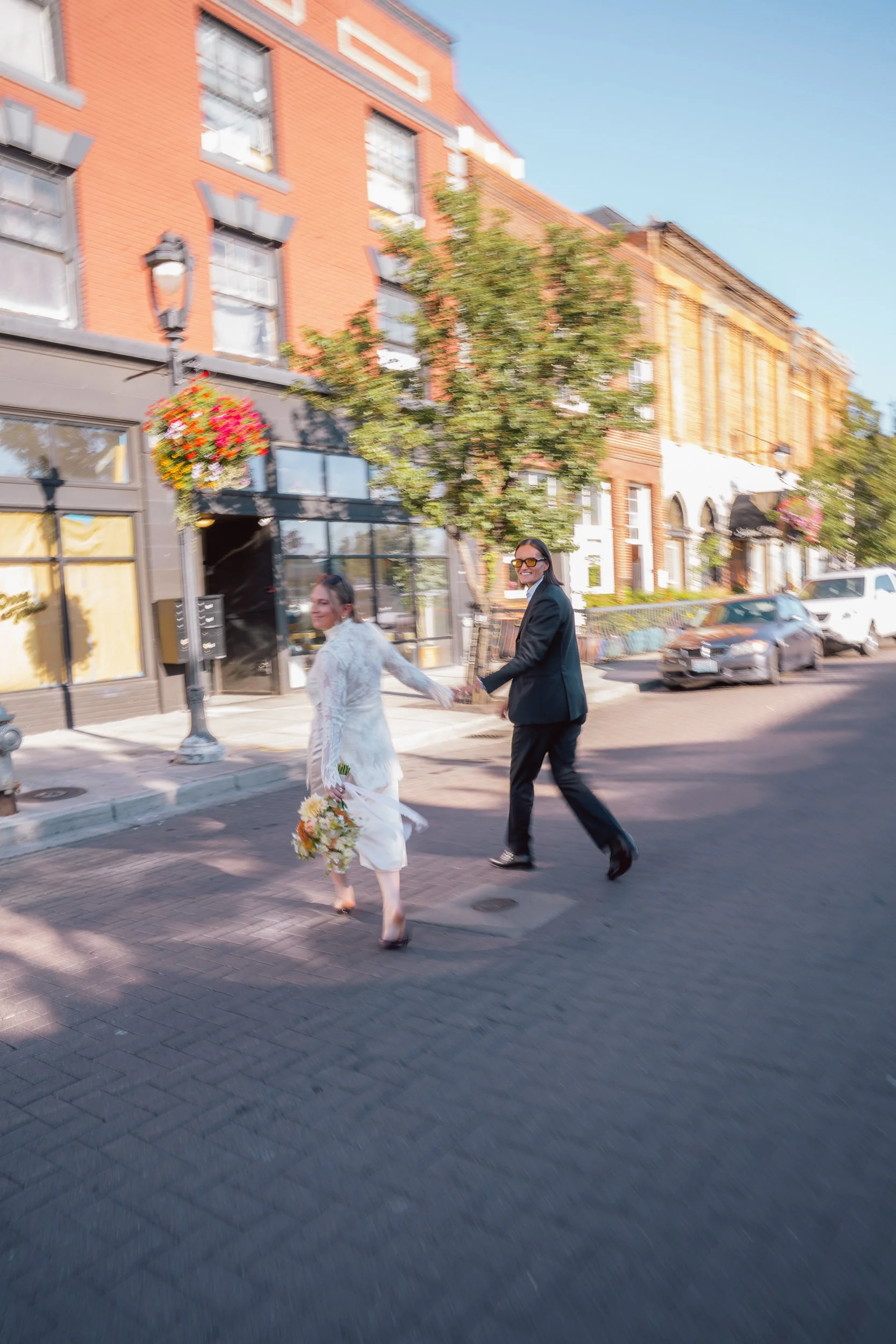 A smiling woman in a black suit and sunglasses holding hands with a woman in a white dress and furry jacket holding a bouquet of flowers, walking across a city street with brick buildings and cars in the background on a sunny day.