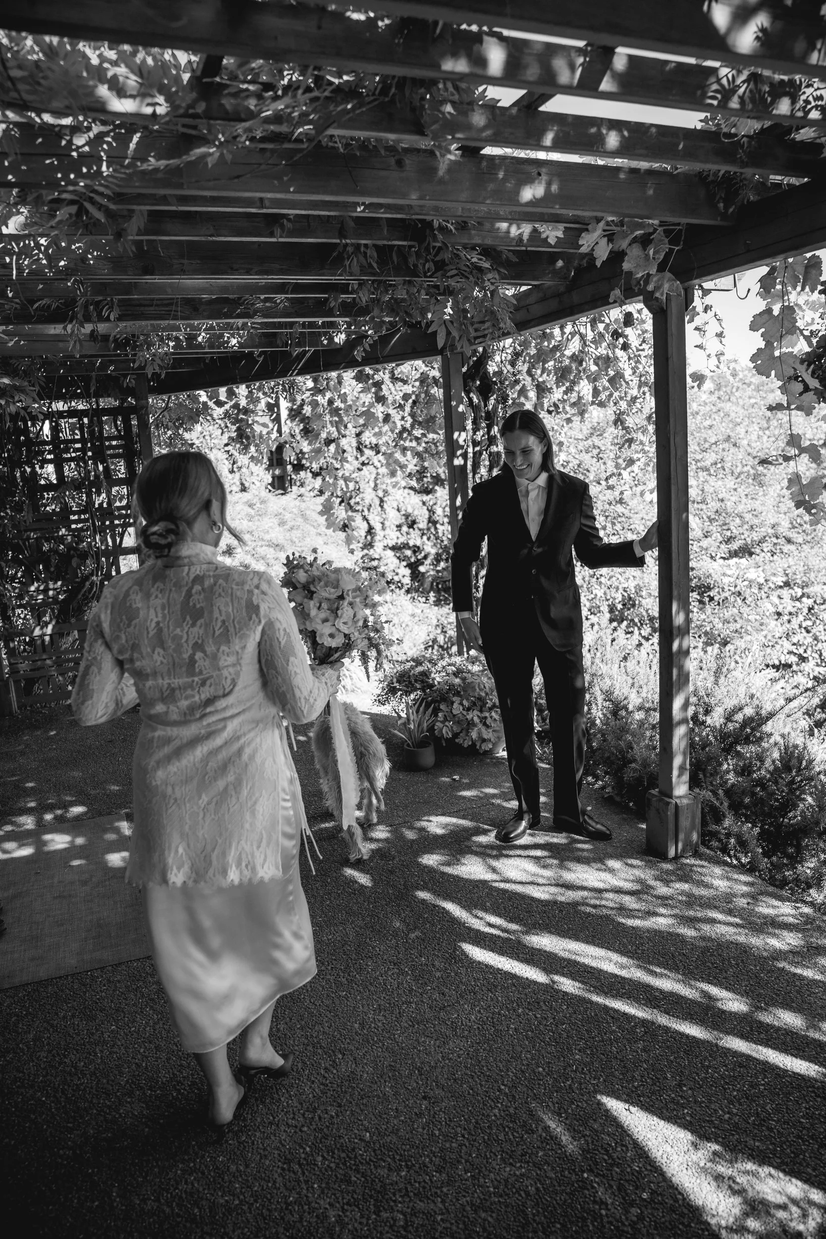 A woman in a lace dress holding a bouquet of flowers and a dog, facing a woman in a suit standing under an outdoor pergola, with trees and sunlight in the background.
