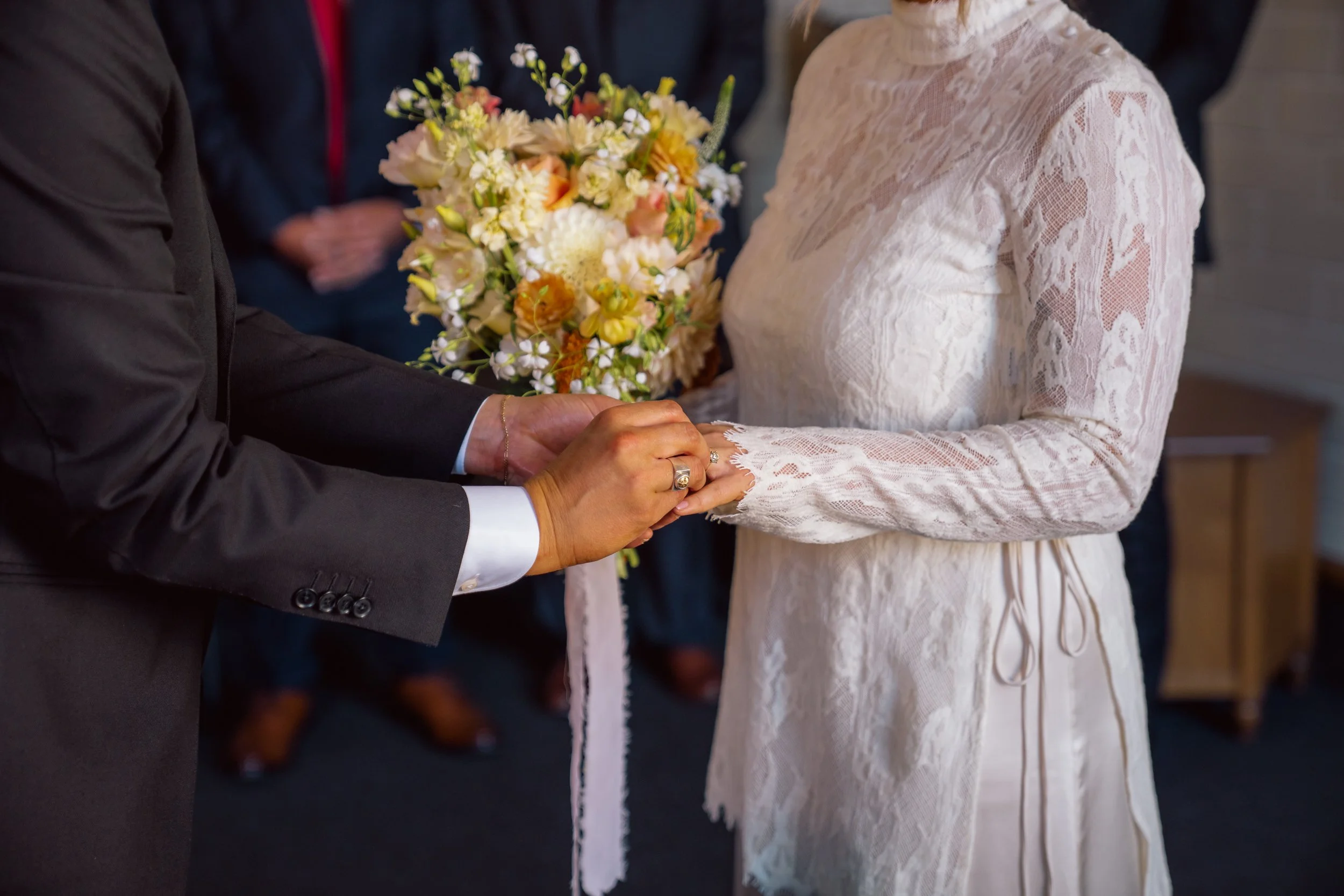 A couple holding hands during a wedding ceremony, with the groom placing a ring on the bride's finger and the bride holding a bouquet of flowers.