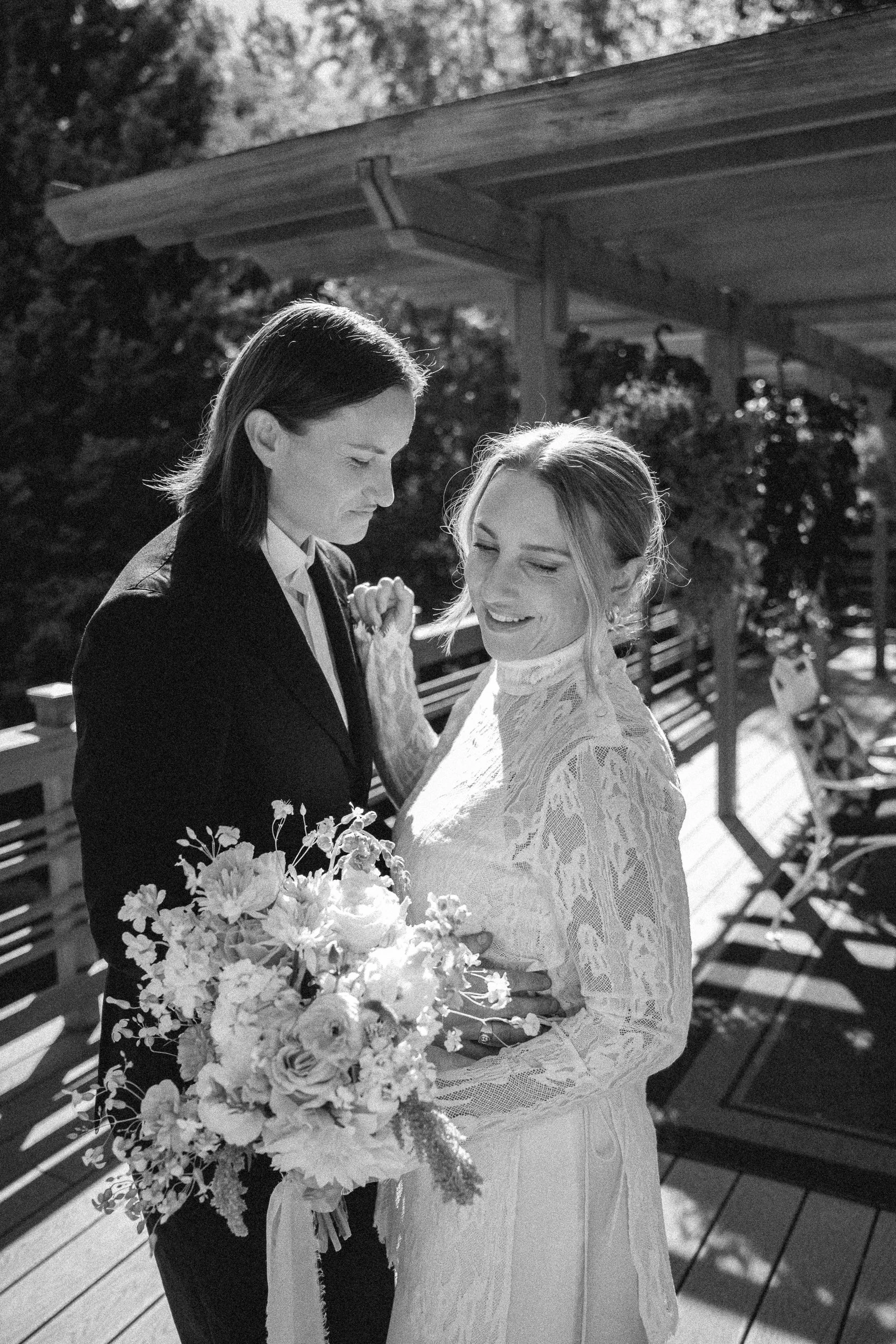 Black and white photo of two women, one in a dark suit and the other in a lace dress, holding hands and smiling while looking at each other on a wooden deck outside, with trees and benches in the background.