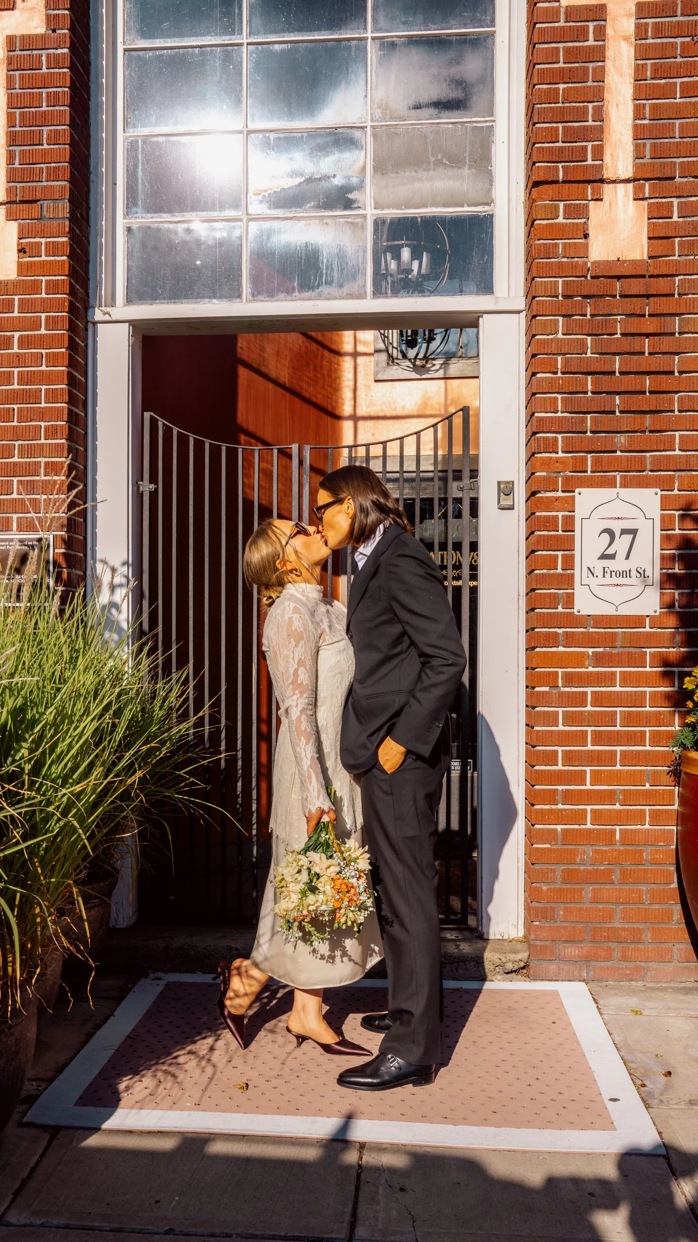 Two women, one in a white dress holding a bouquet and the other in a black suit, sharing a kiss in front of a house entrance.