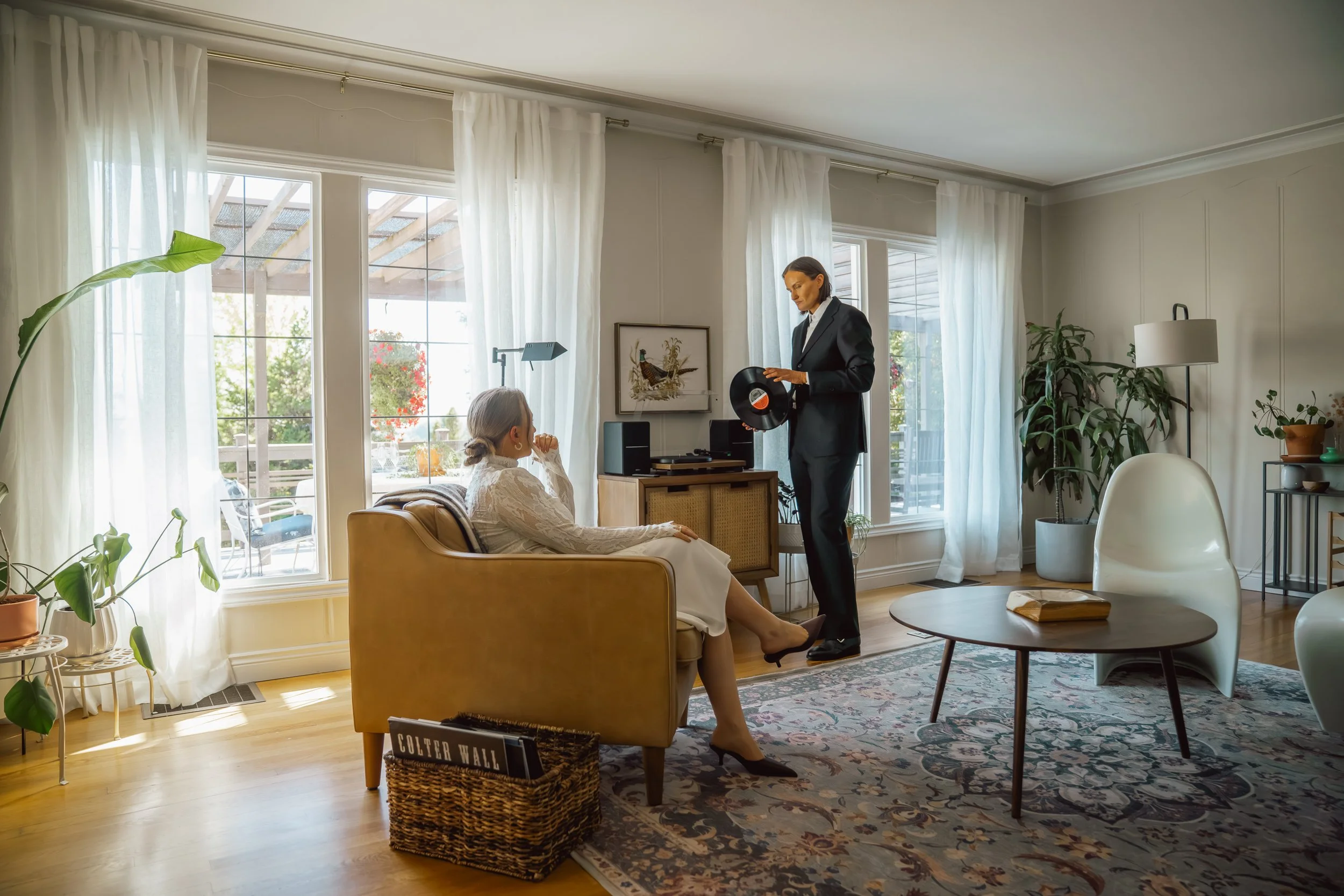 A woman in a white dress sitting on a yellow sofa listening to a man in a black suit holding a vinyl record inside a bright living room with large windows, white curtains, and various plants.